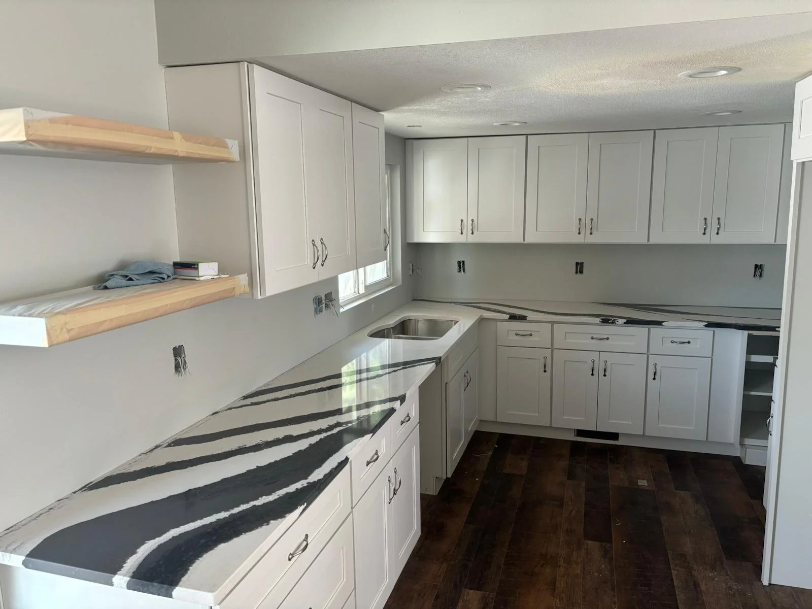 Kitchen with white cabinets, dark wood flooring, and black and white patterned marble countertop. Shelves, no appliances, uninstalled electrical outlets, and a window above the sink.