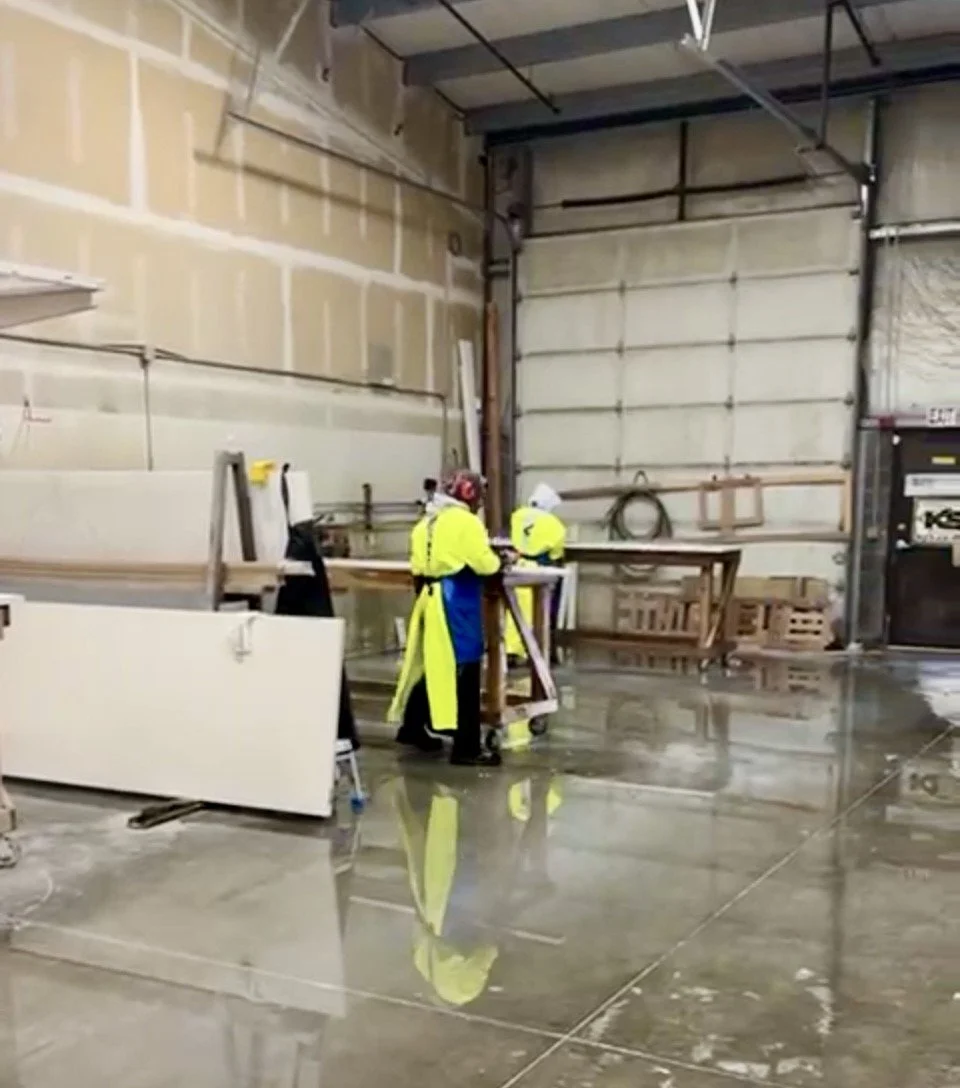 Two workers in yellow safety jackets and white hard hats working in an industrial warehouse with a wet concrete floor.