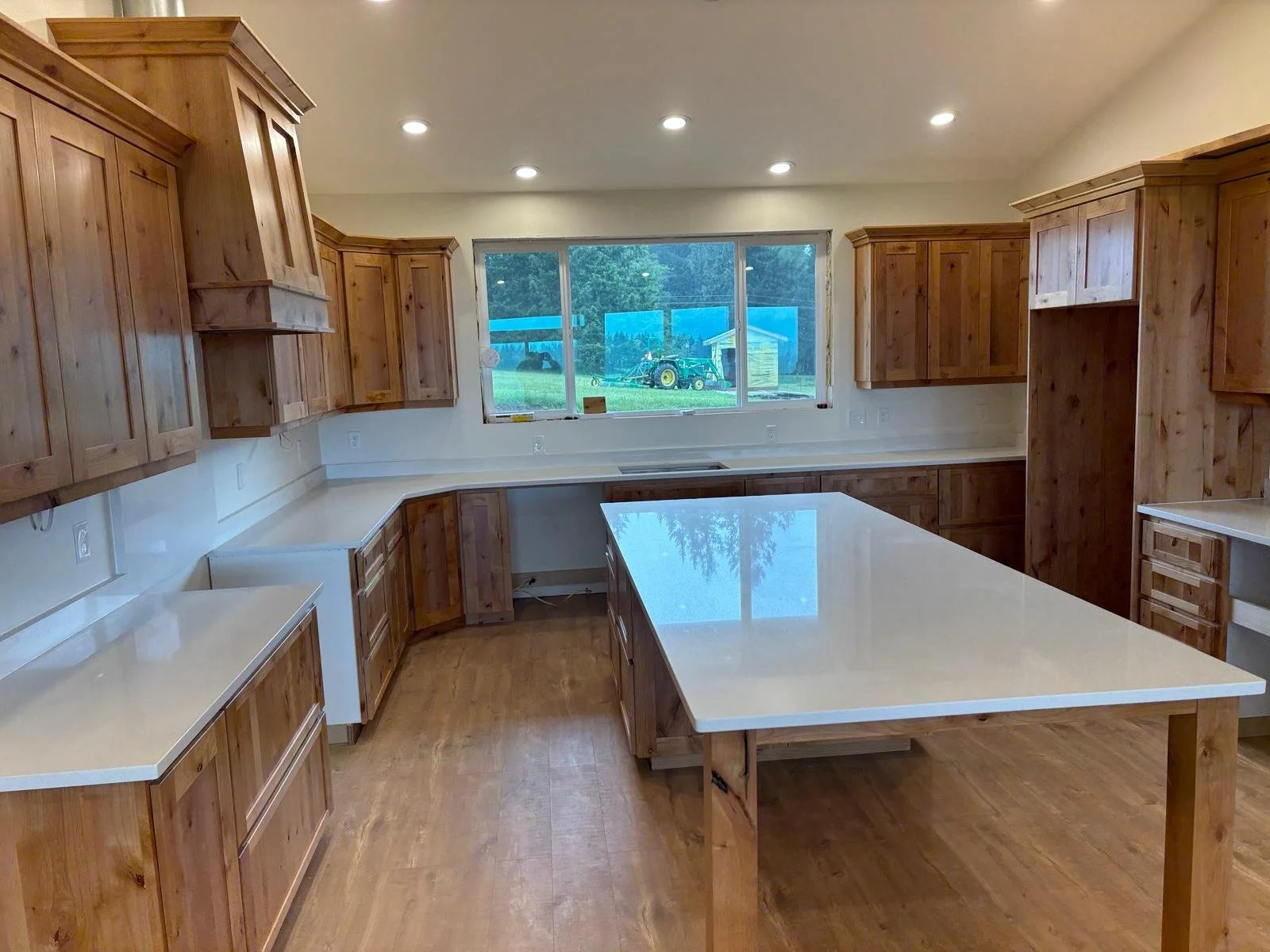 A large kitchen with wooden cabinets, white countertops, and a kitchen island in the center. There is a window overlooking a yard with outdoor play equipment and a tractor.