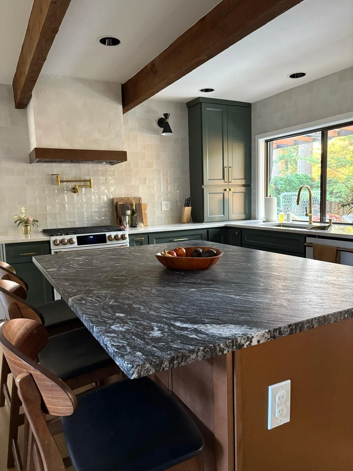 Modern kitchen with dark green cabinets, a large marble kitchen island, and a window overlooking a backyard with green trees.