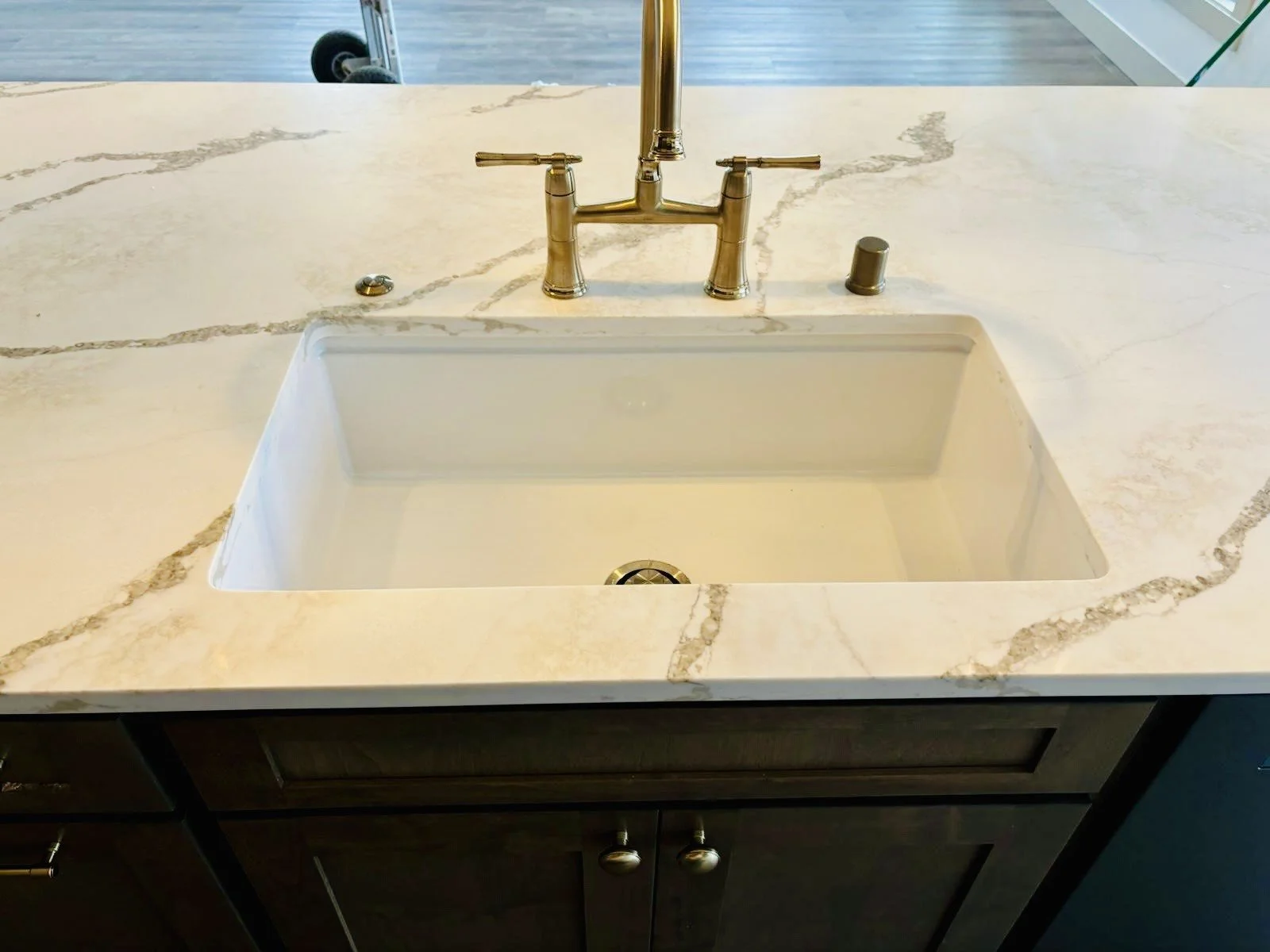 Kitchen sink with a gold-colored faucet, surrounded by a light-colored marble countertop, with a dark wooden cabinet underneath.