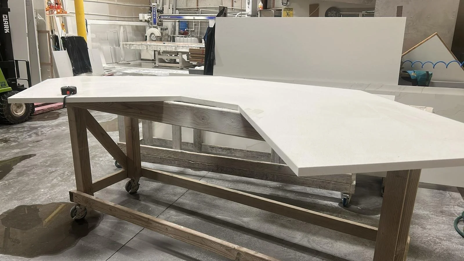 A large work table with a white top, adjustable on a wooden frame with casters in a workshop. The table is located in a furniture workshop with tools and materials in the background.