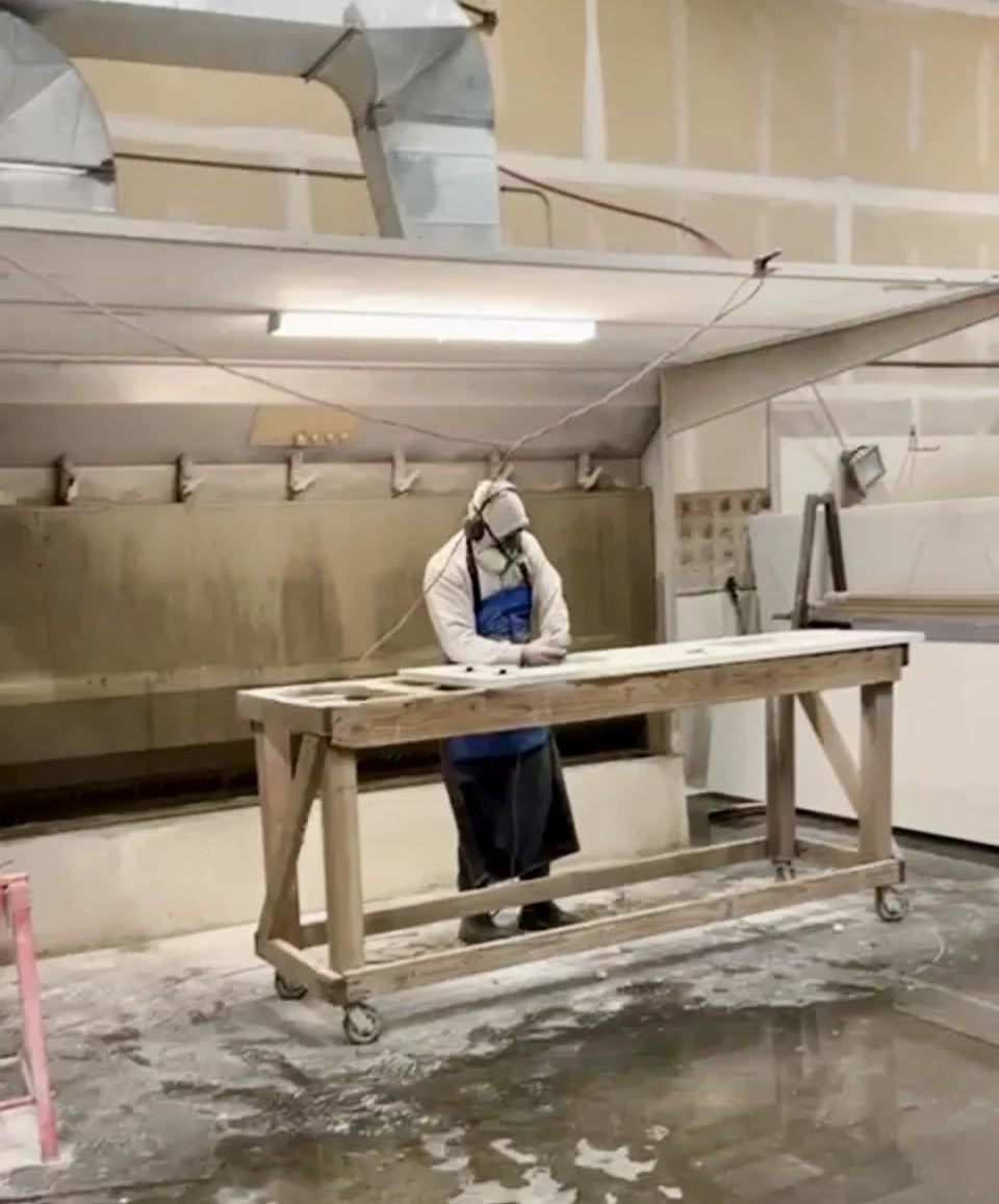 A person wearing protective gear is working in a workshop, spraying or painting a large flat piece of material on a wooden worktable. The workspace has industrial equipment and a beige wall in the background.
