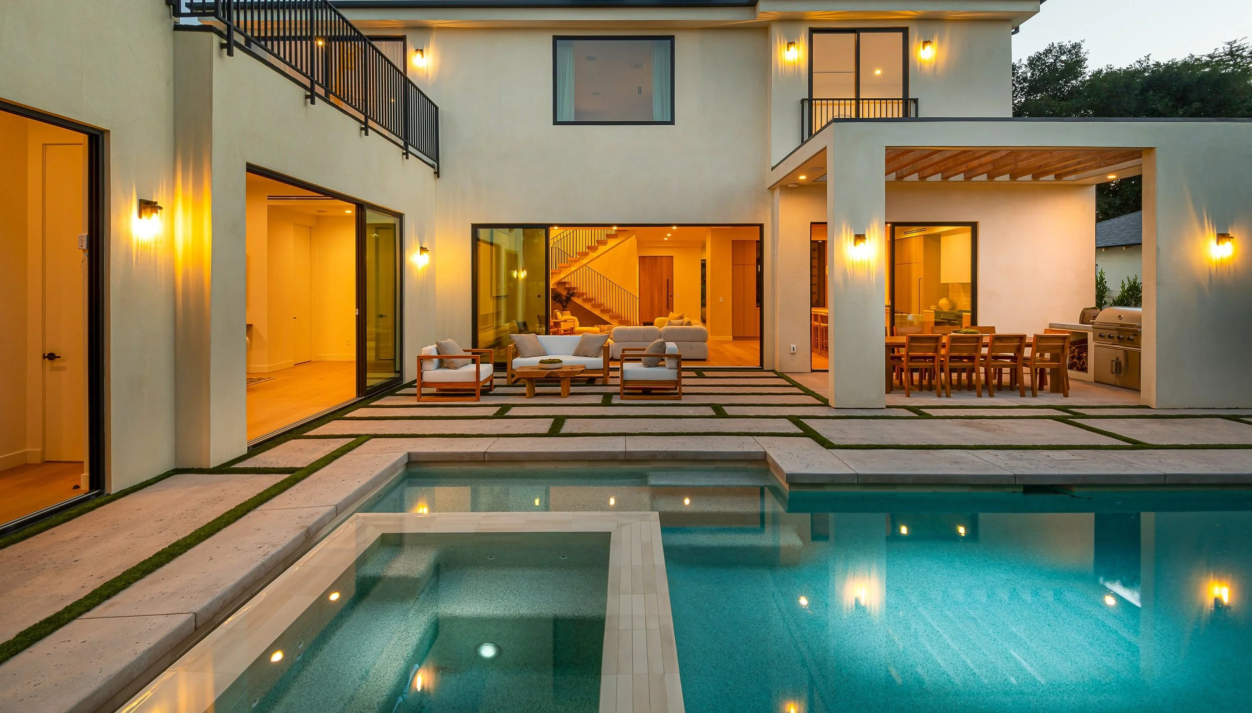 Modern house with a backyard pool, outdoor seating, and an outdoor kitchen area at dusk, with lights illuminating the patio.