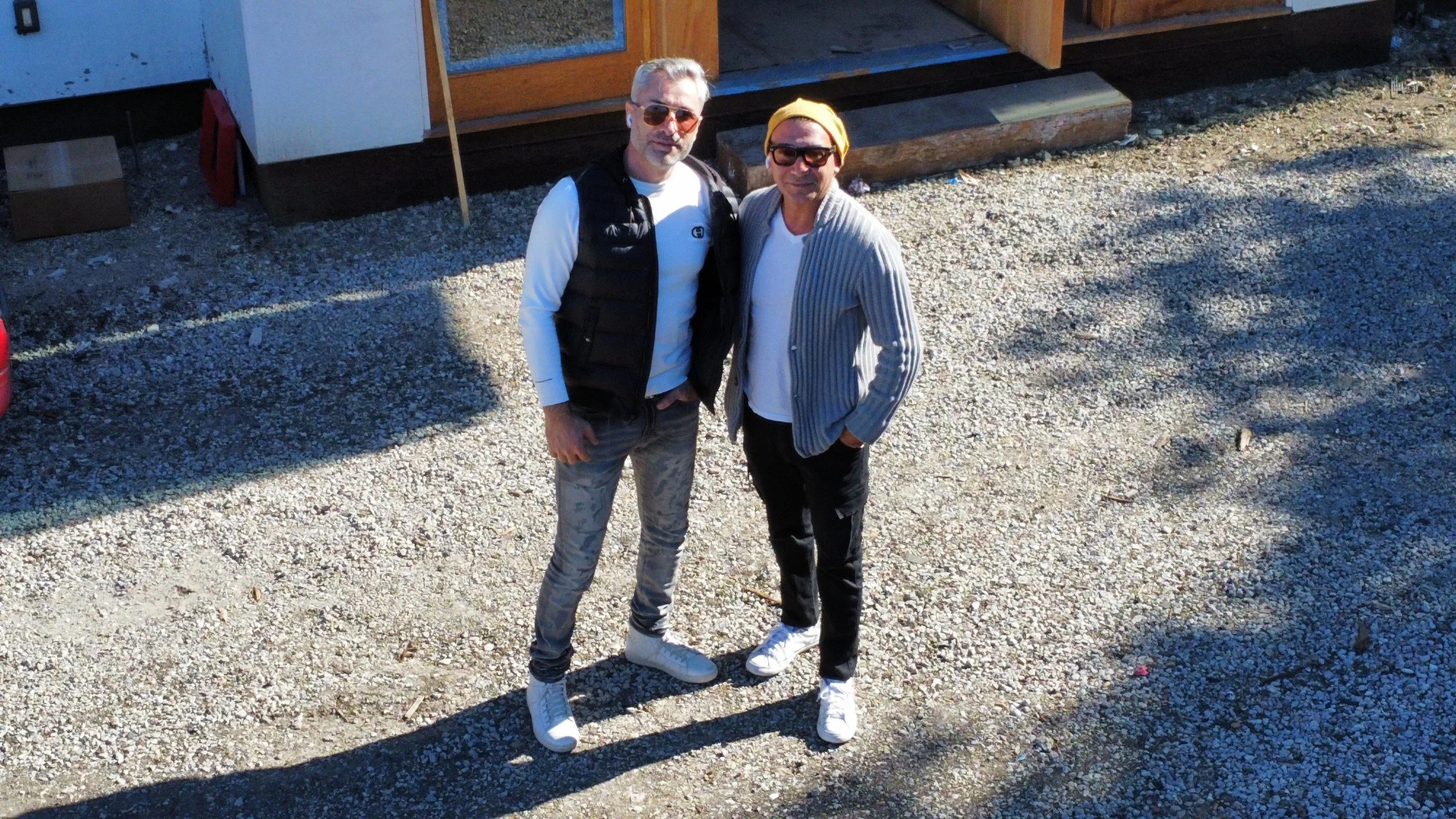Two men standing on a gravel surface outside, posing for a photo. One man has white hair, sunglasses, wearing a black vest over a white shirt, and camouflage pants. The other man has a yellow beanie, sunglasses, a gray striped blazer, and black pants.
