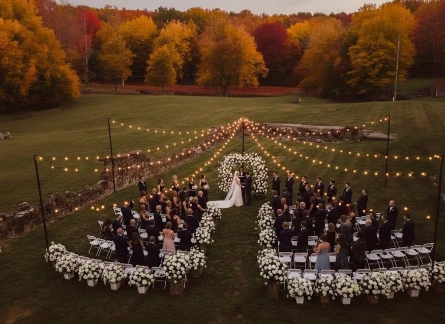 Outdoor wedding ceremony with a bride and groom exchanging vows under a floral arch, surrounded by seated guests, white flower arrangements, and string lights hanging overhead on a grassy field during dusk, with autumn trees in the background.