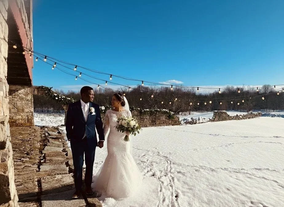 A bride and groom holding hands and walking in a snowy outdoor setting during a wedding photoshoot, with string lights overhead, a stone building to the left, and a distant wooded landscape under a clear blue sky.