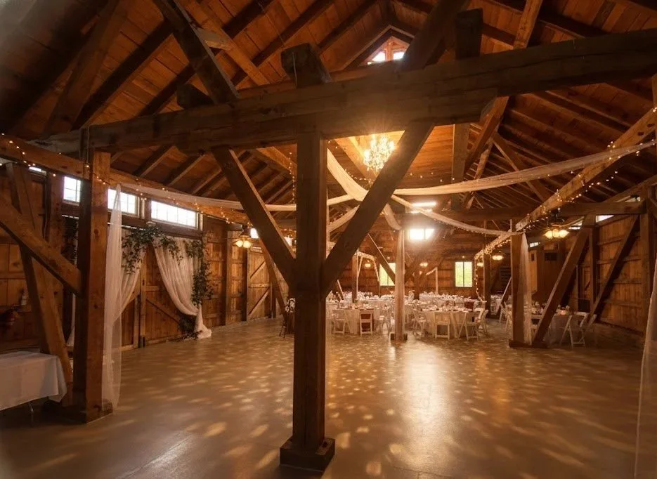 A decorated rustic barn reception space with round tables, white chairs, draped fabric, and string lights.
