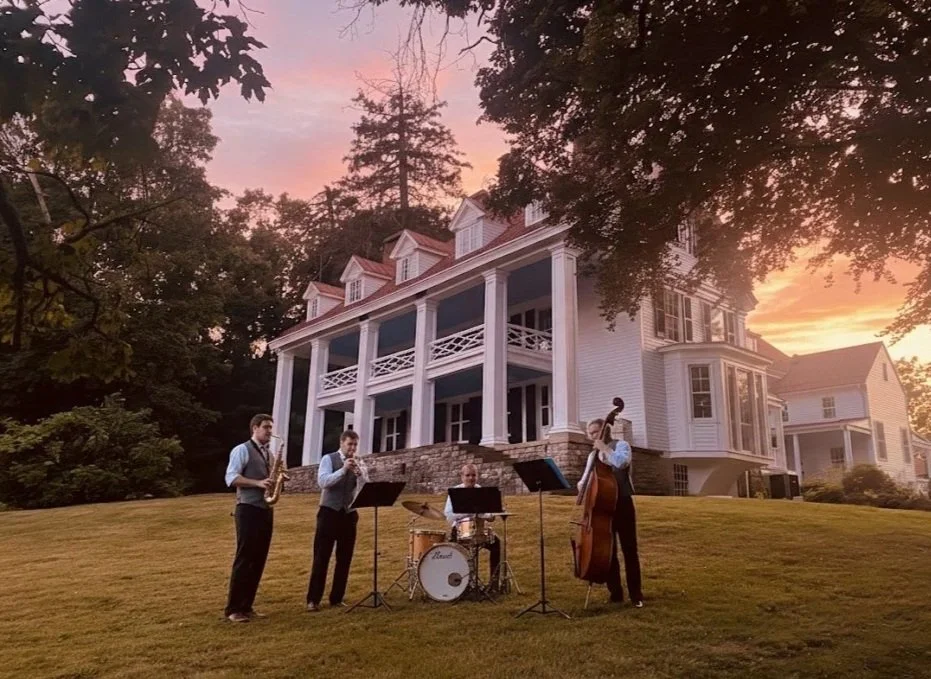 A jazz band performing outside on a grassy lawn in front of a large, white, Greek revival-style house during sunset. The band consists of four musicians playing saxophone, trumpet, drums, and double bass.