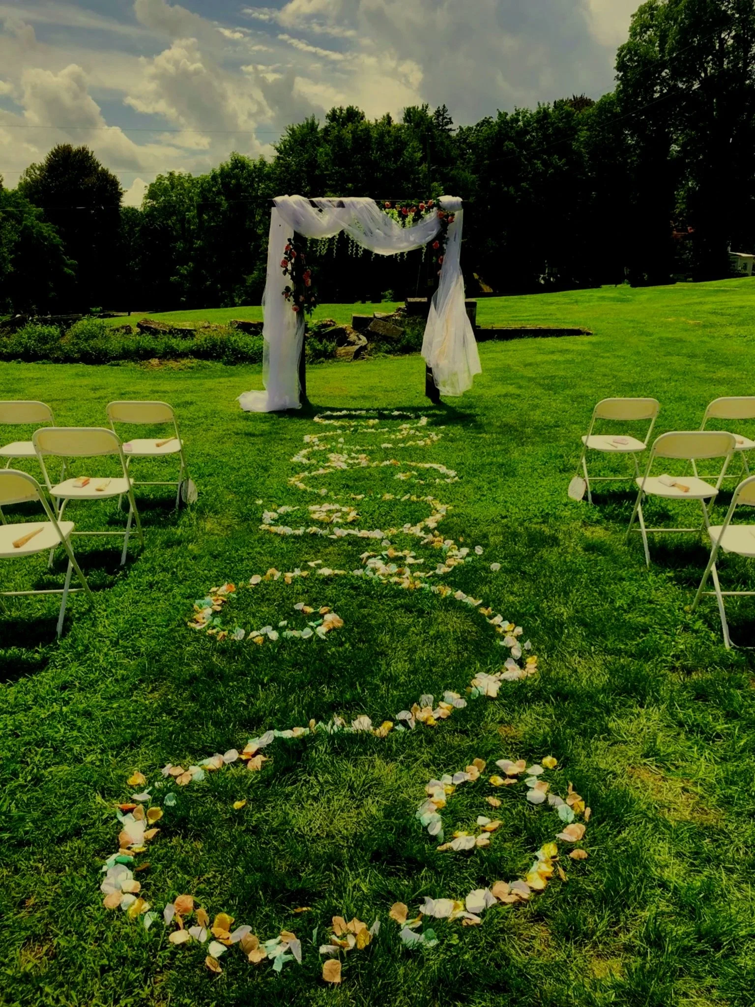 Outdoor wedding ceremony setup with white chairs on either side of a rose petal aisle leading to a decorated arch, surrounded by green grass and trees under a partly cloudy sky.