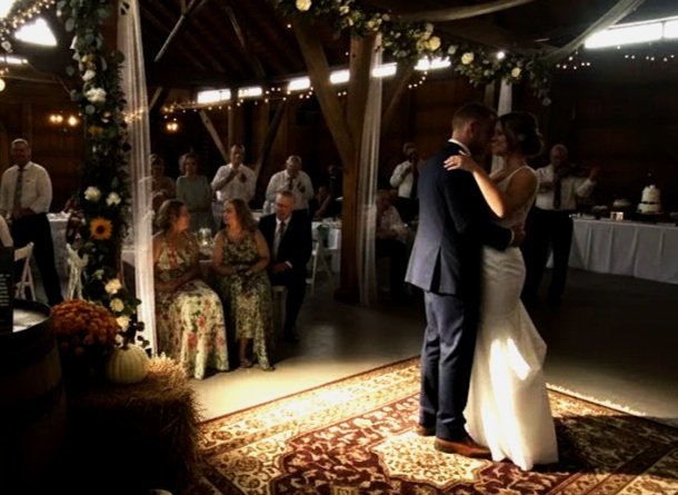 A bride and groom are sharing their first dance under a decorated flower arch at a wedding reception inside a barn. Guests are seated at tables, watching the couple. The decor includes pumpkins, flowers, and fairy lights, with a rustic theme.
