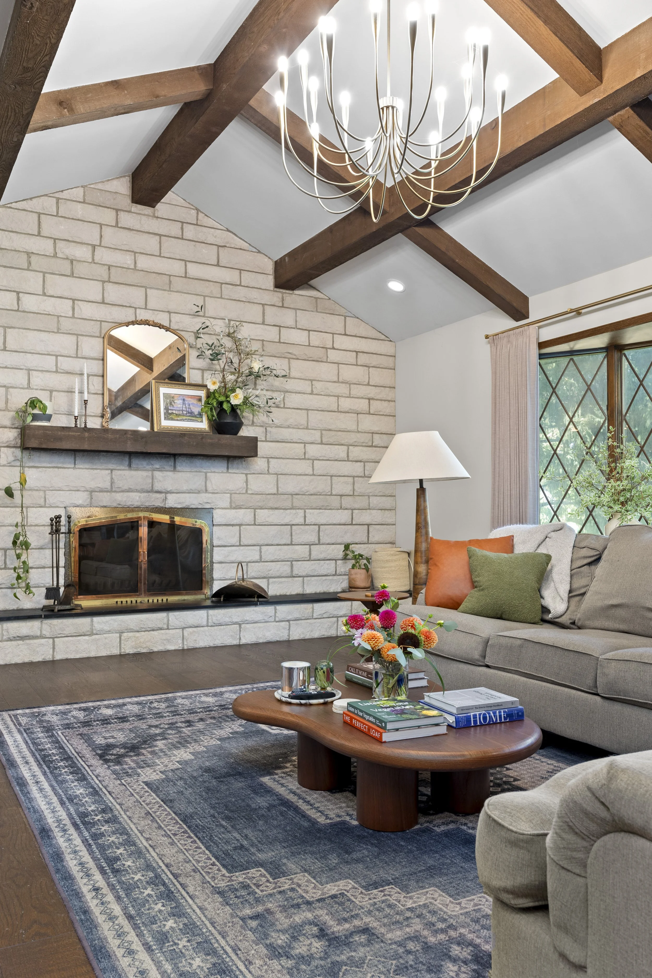 Living room with a beige sofa, colorful pillows, and a wooden coffee table with books, flowers, and a tray. A patterned blue rug covers the hardwood floor. There is a white brick fireplace with a wooden mantel decorated with a mirror, framed picture,