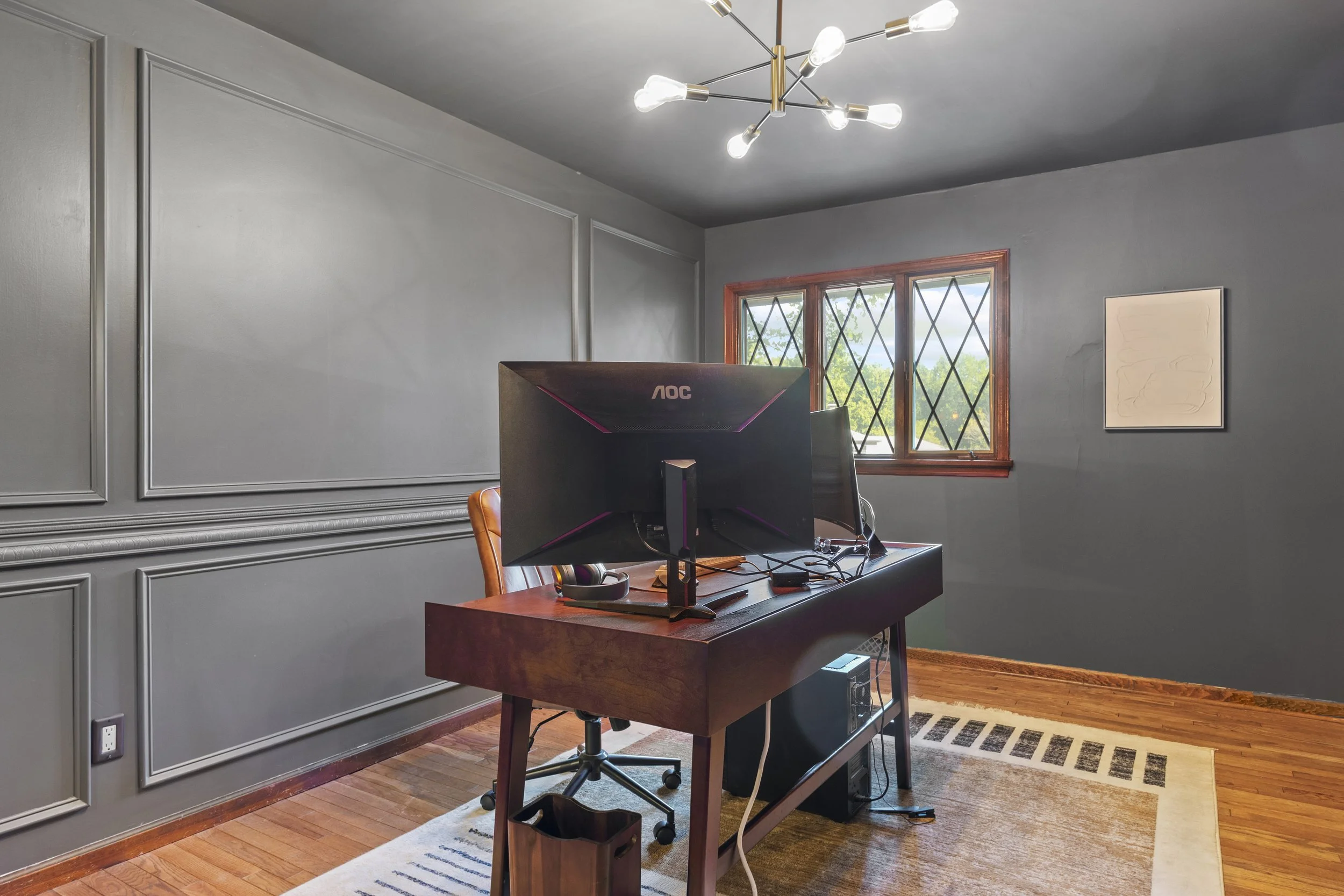 A home office with a wooden desk holding a computer monitor, a keyboard, and a chair. The room has gray walls, decorative wall molding, a window with diamond-shaped panes, a modern ceiling light fixture with exposed bulbs, and a colorful rug on hardw