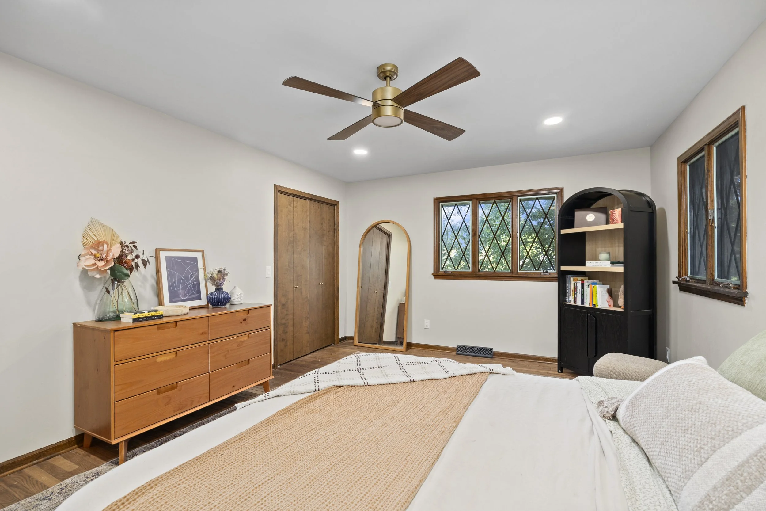 A cozy bedroom with a white bed, a wooden dresser with vases and artwork, a black bookshelf, a mirror, window with diamond-shaped grids, and a ceiling fan.
