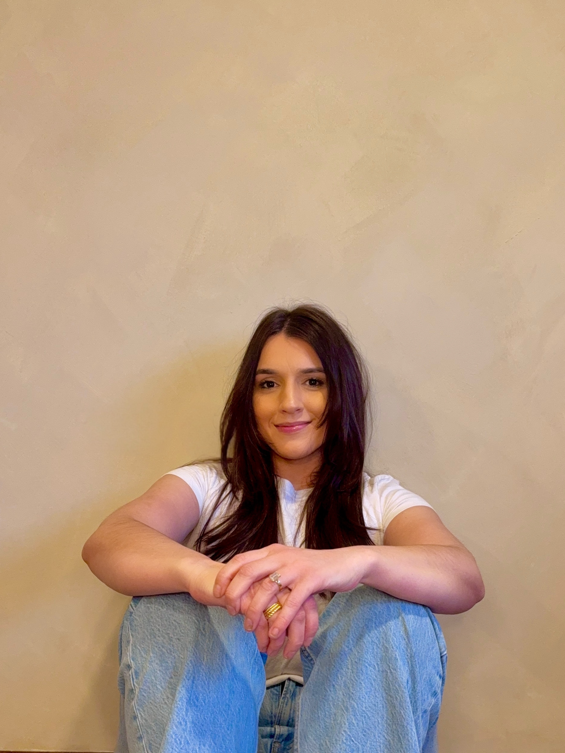 A woman sitting on the floor against a plain beige wall, smiling at the camera, wearing a white T-shirt and blue jeans, with her hands clasped together.