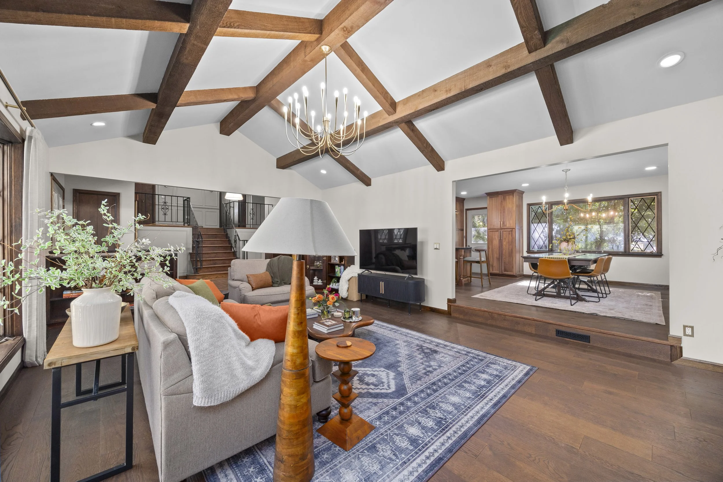 Living room with vaulted ceiling, wooden beams, modern chandelier, white walls, brown hardwood floors, with furniture including a gray sofa with pillows, a wooden side table, a white lamp, a black TV stand with a TV, dining table with four chairs, la
