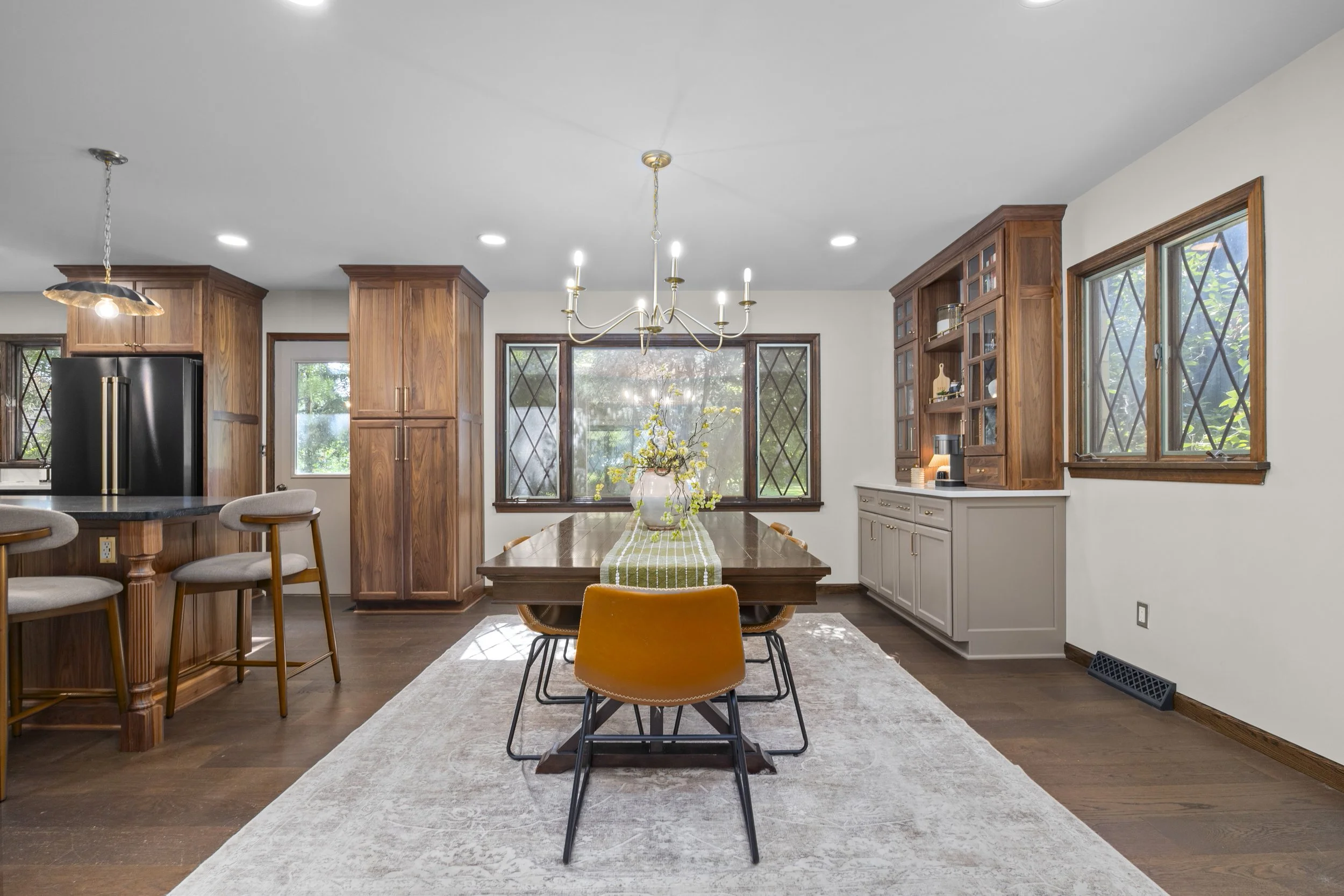 A dining area with a wooden table, yellow chairs, and a vase with yellow flowers. Large windows with diamond-shaped panes let in natural light. Wooden cabinets and a chandelier hang from the ceiling.