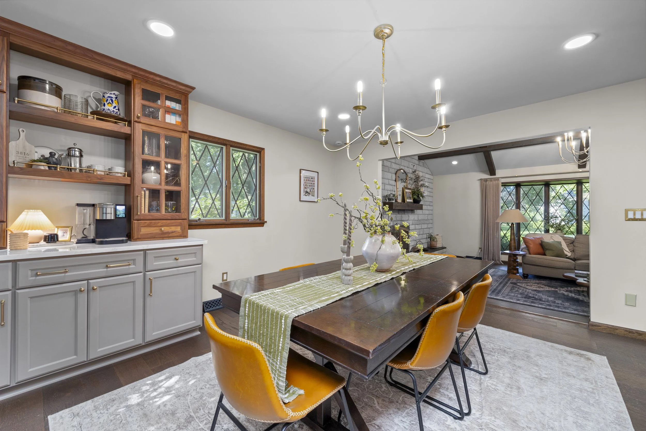 Dining room with a wooden table, yellow chairs, a vase with flowers, and a view into the living room with a sofa and windows.