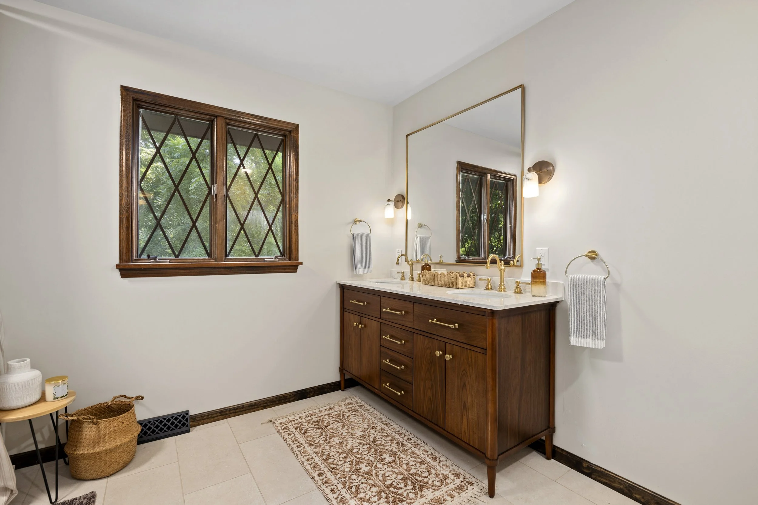 Bathroom with white walls, two windows with diamond-shaped wooden frames, a large mirror, wall-mounted lights, a wooden vanity with brass handles, a marble countertop, and decorative items including a soap dispenser, a towel, and a decorative basket.
