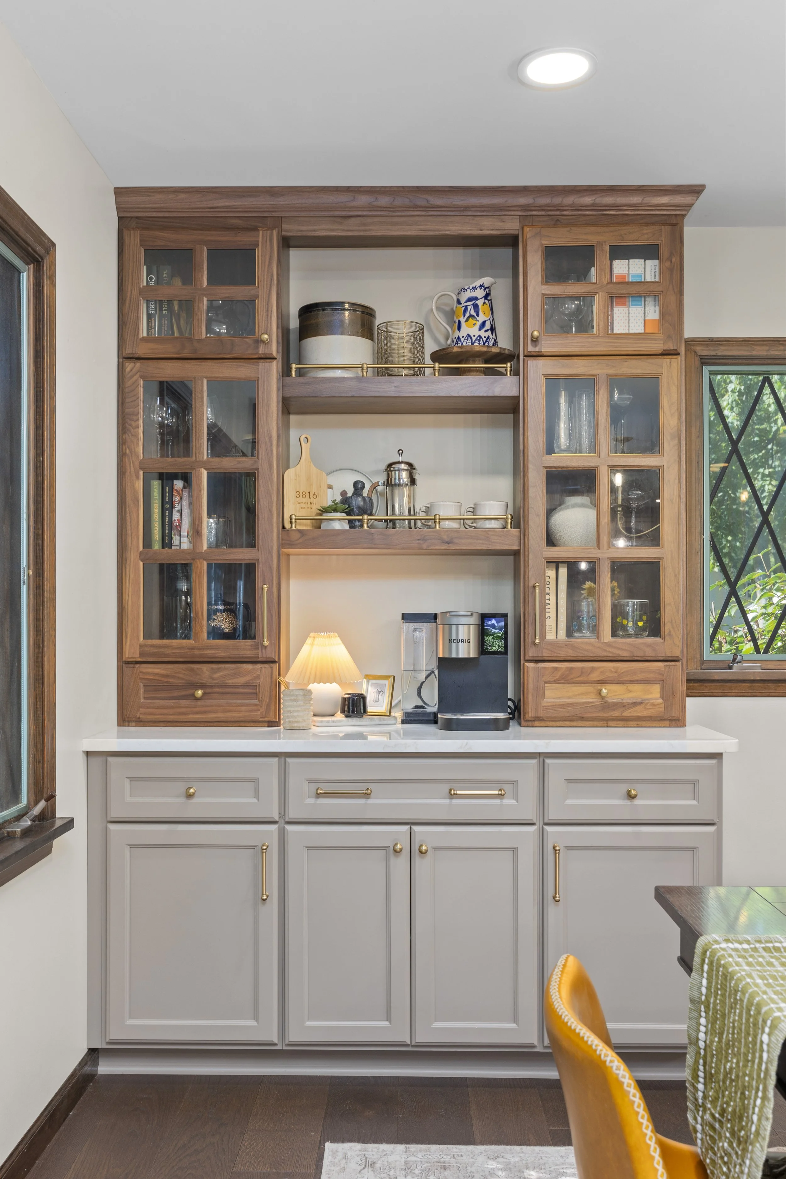 A kitchen cabinet with glass-paneled doors and open shelves, containing dishes, books, and decorative items, with a white counter below and a window to the right showing greenery outside.