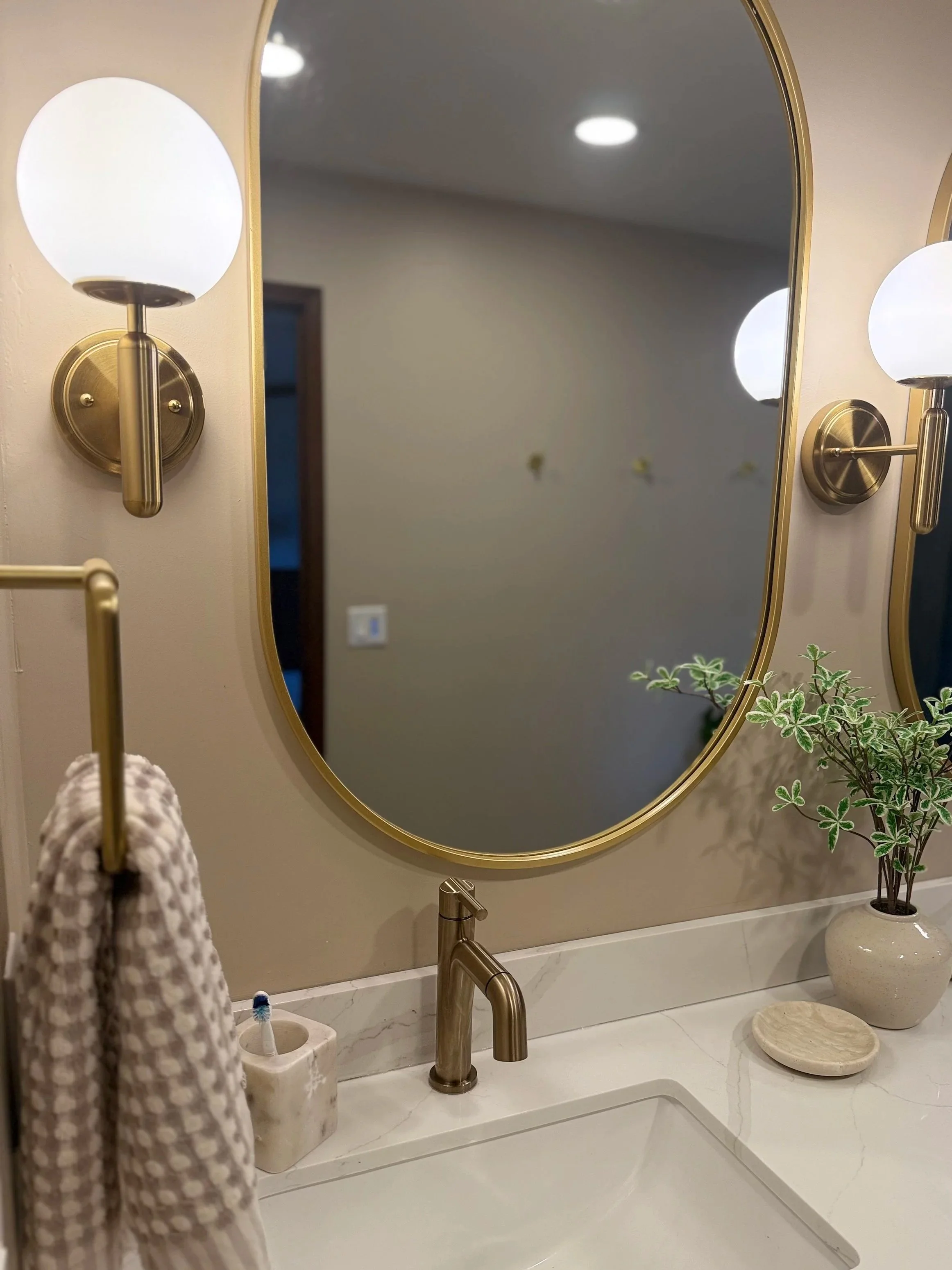 Bathroom vanity with a large oval mirror, two wall-mounted light fixtures, a beige towel hanging on a ring, a toothbrush holder with a toothbrush, a potted plant, and a decorative plate or stone on the countertop.