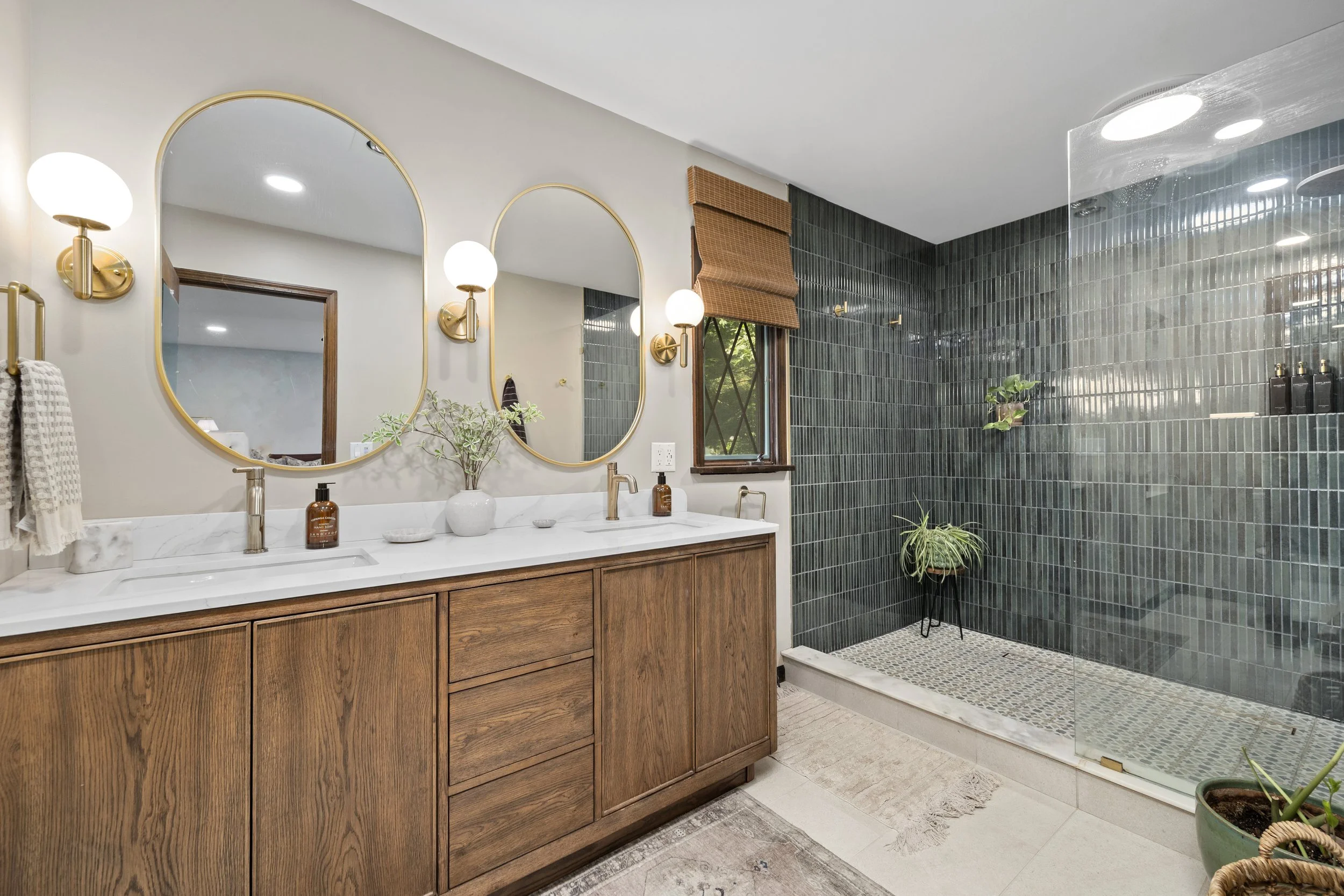 A modern bathroom with a wooden vanity, oval mirrors with golden frames, white marble countertop, and gold wall sconces. There are plants on the counter and in the shower area, which has dark green tile walls and a glass enclosure.