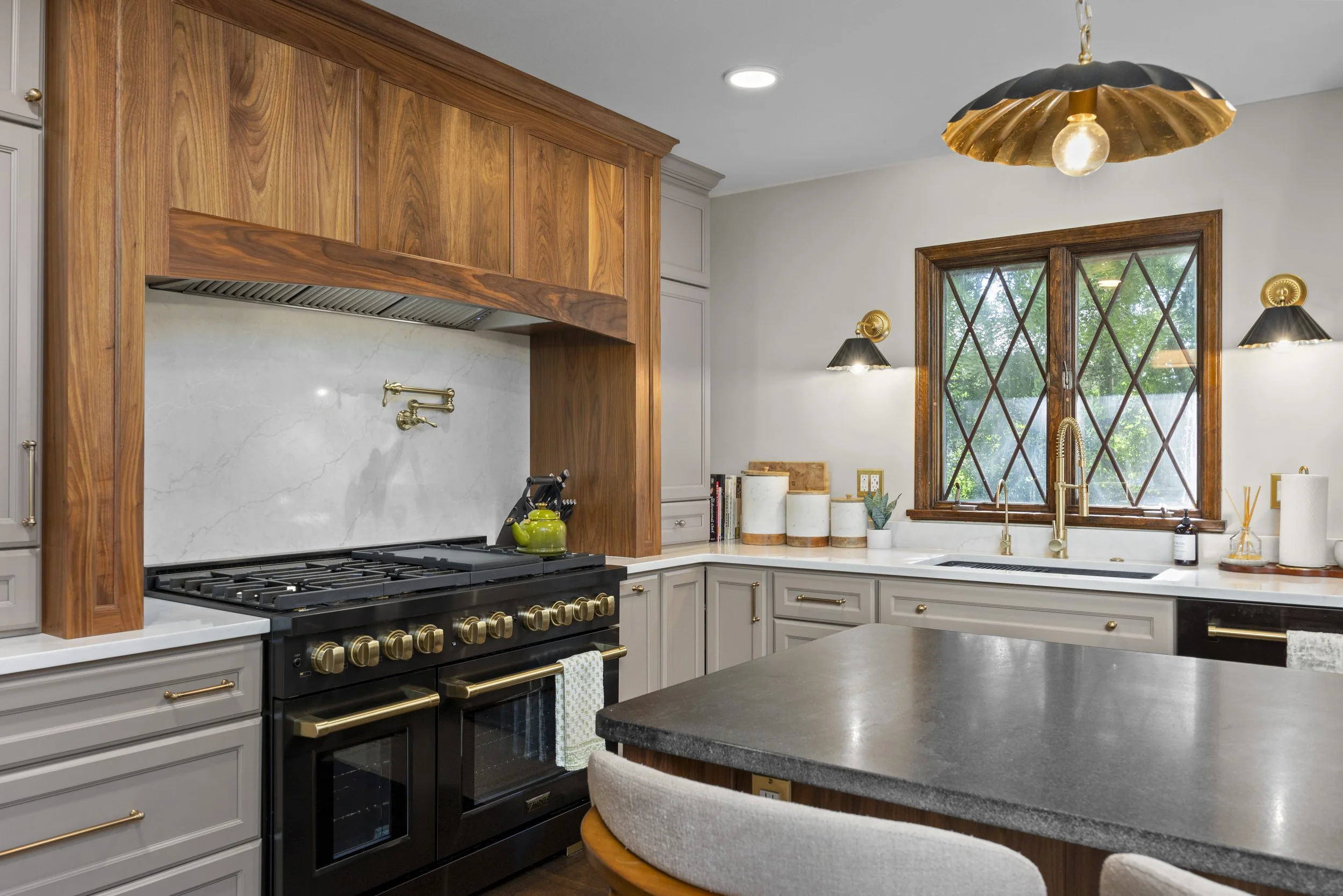 Kitchen with white cabinets, black stove with gold knobs, wooden range hood, marble backsplash, large window, gold fixtures, and a gray countertop
