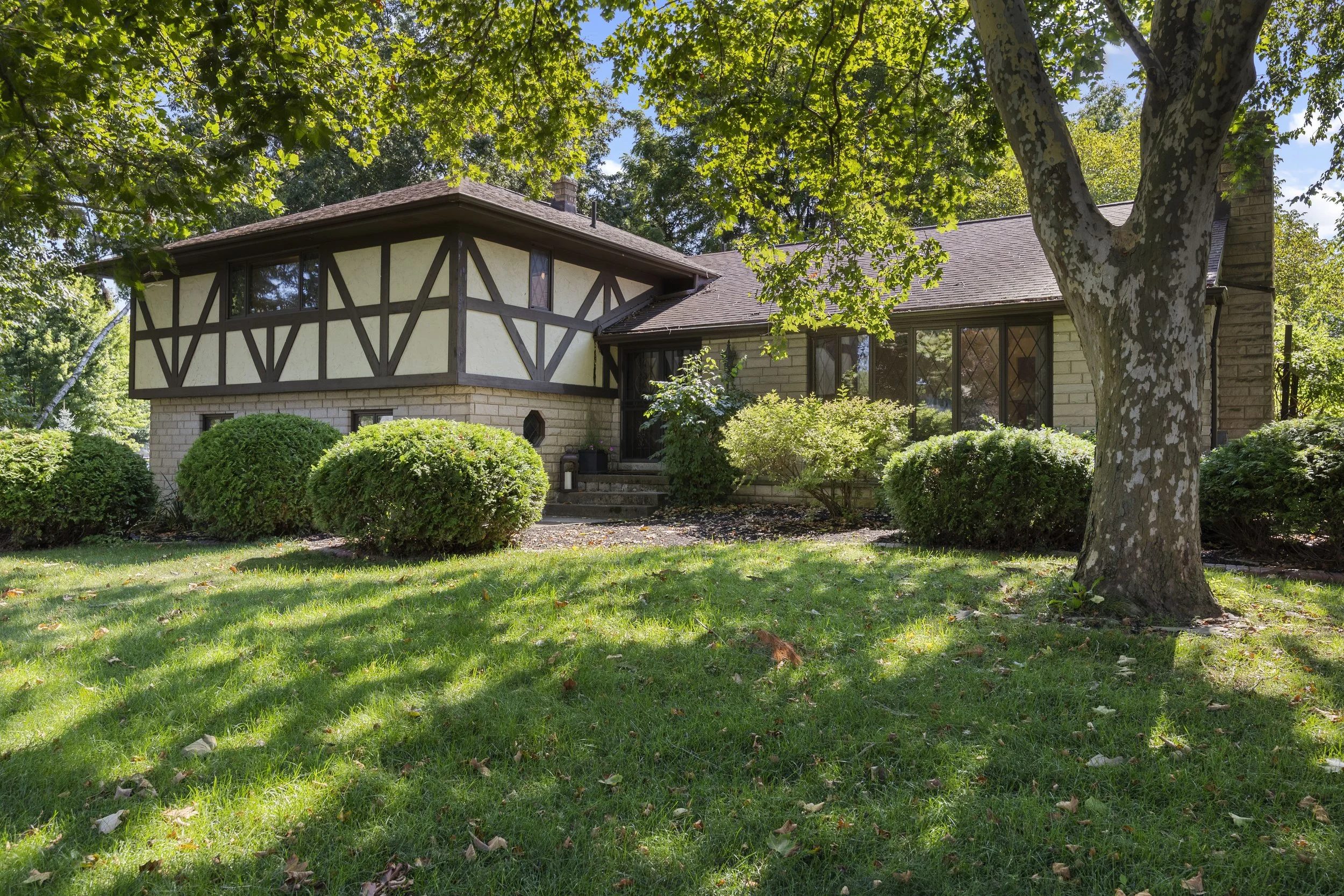 A house with a Tudor-style exterior, large windows, brick and stucco walls, surrounded by lush greenery and trees, on a sunny day.