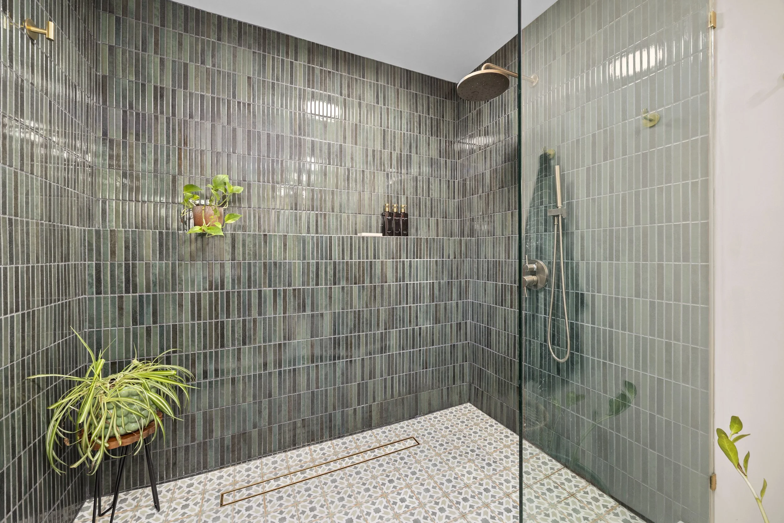 Modern shower with tiled green walls, plant decor, and patterned floor tiles, featuring a rainfall showerhead, a hand-held shower, and black bottles.