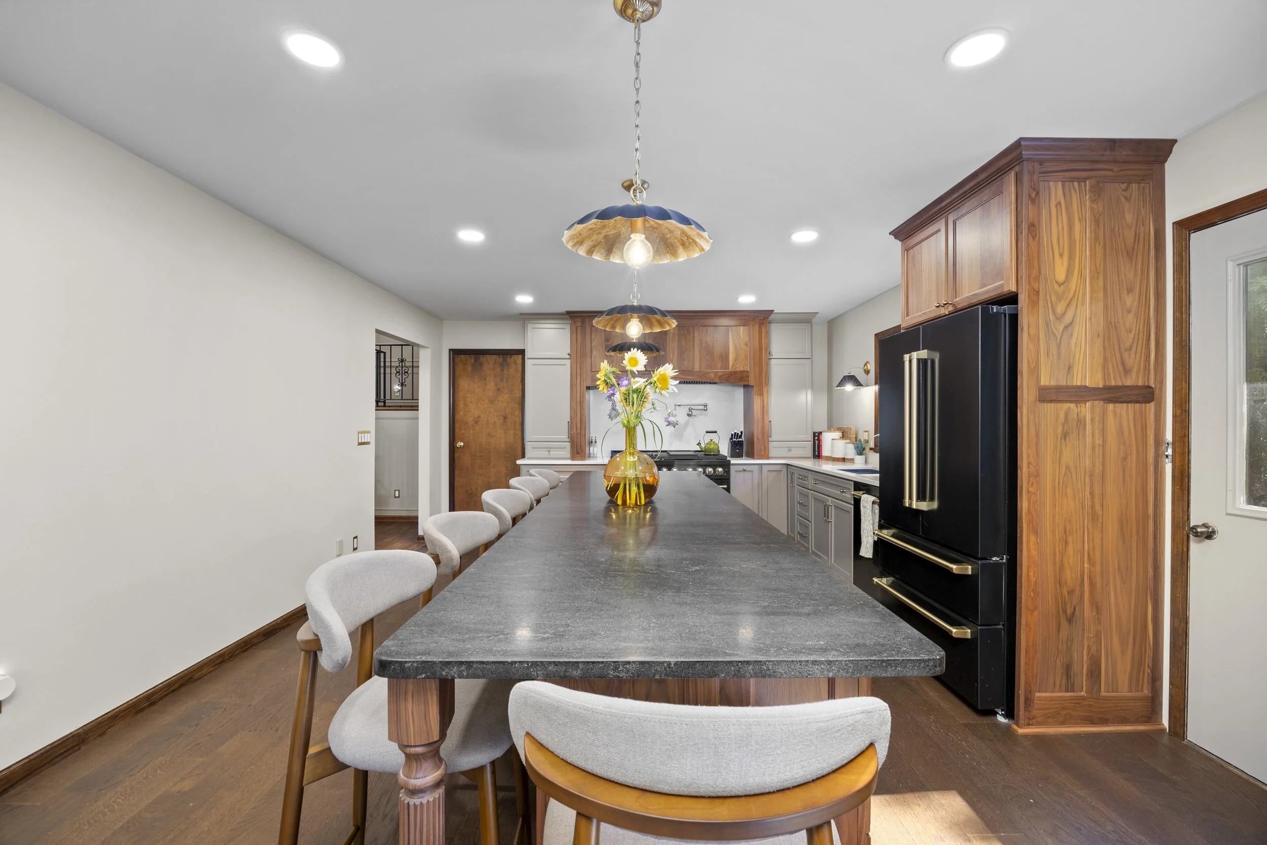 Kitchen with wooden cabinetry, black refrigerator, gray countertop island, white walls, hardwood floors, and decorative pendant lighting with flowers in a yellow vase.