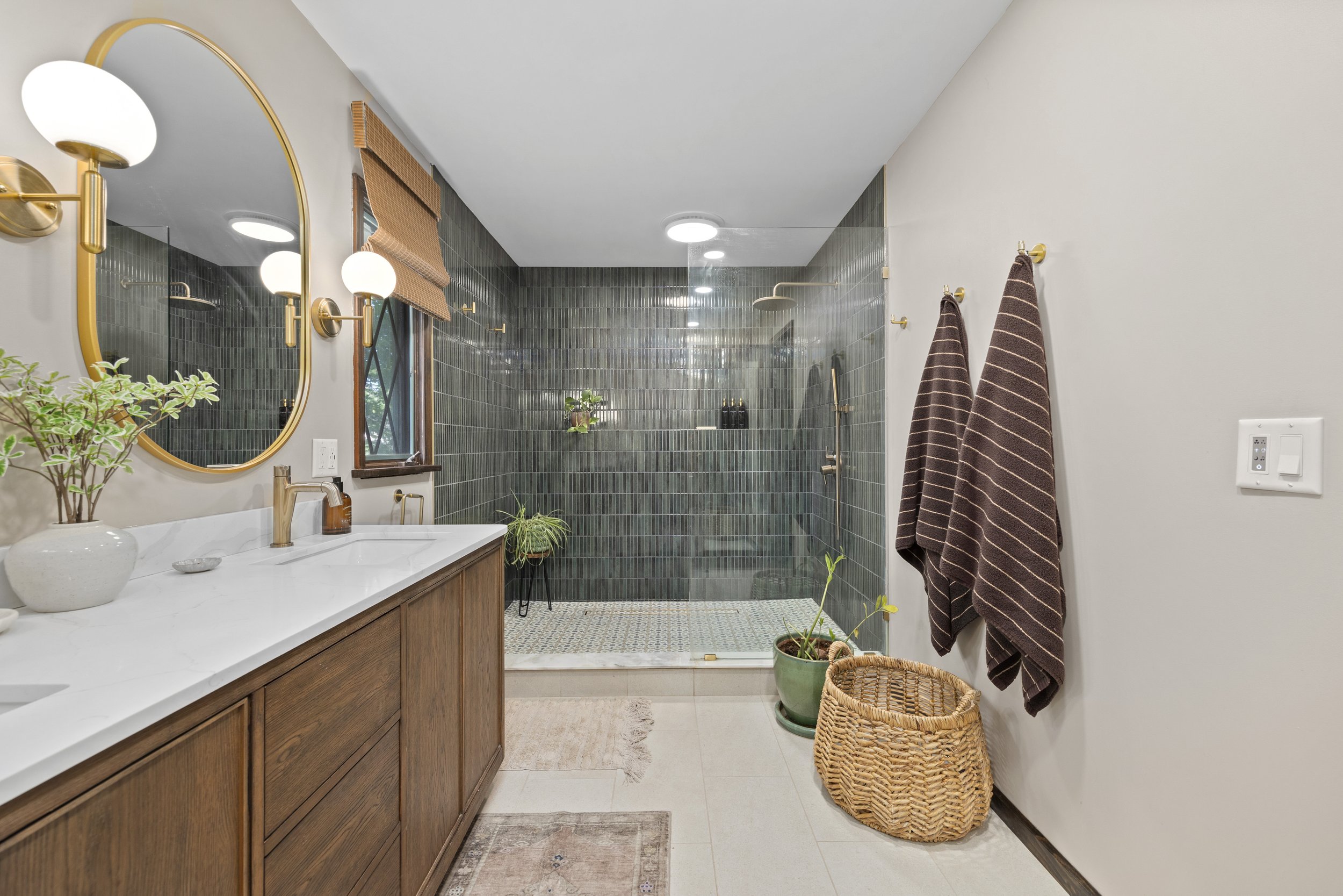 A modern bathroom featuring a white vanity with a marble countertop, gold fixtures, a large round mirror, and wall-mounted light fixtures. There are two brown towels hanging on gold hooks, a wicker basket, and various potted plants. The shower area h