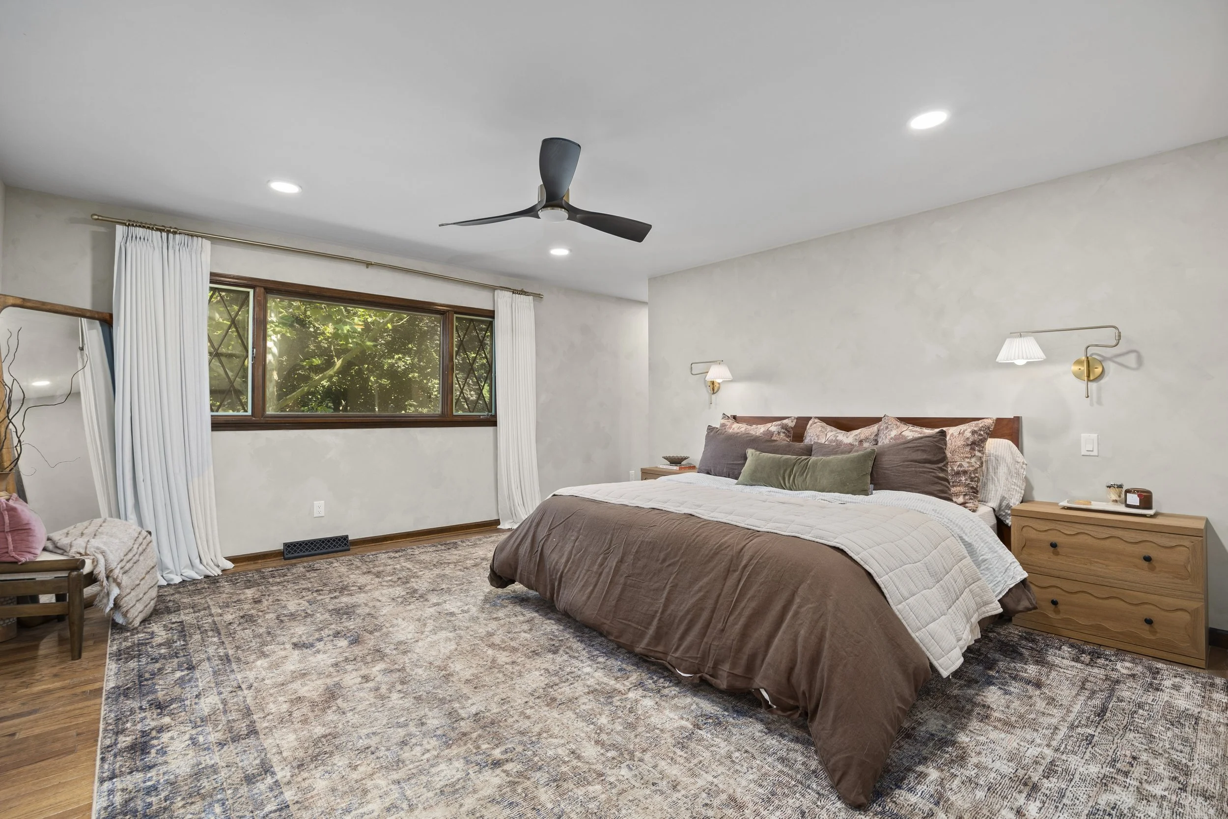 A bedroom with a large bed, wooden nightstands, wall-mounted lamps, a window with white curtains, a ceiling fan, and a patterned area rug.