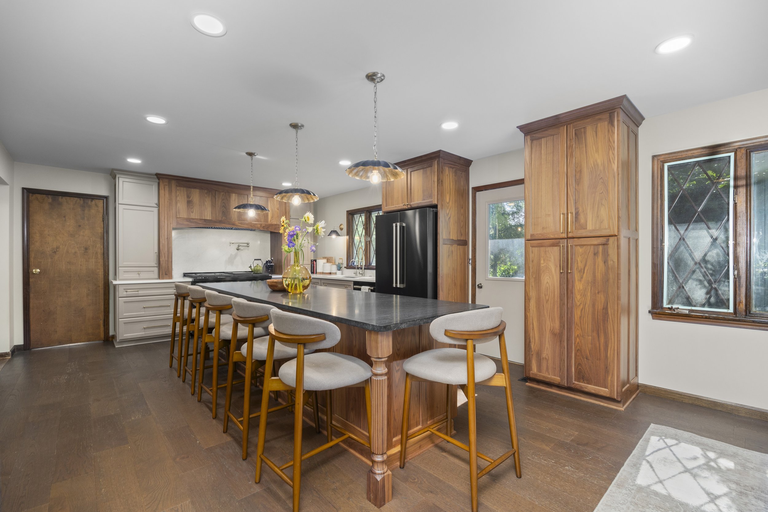 Modern kitchen with wooden cabinets, a kitchen island with beige chairs, a vase with flowers, and stainless steel appliances.