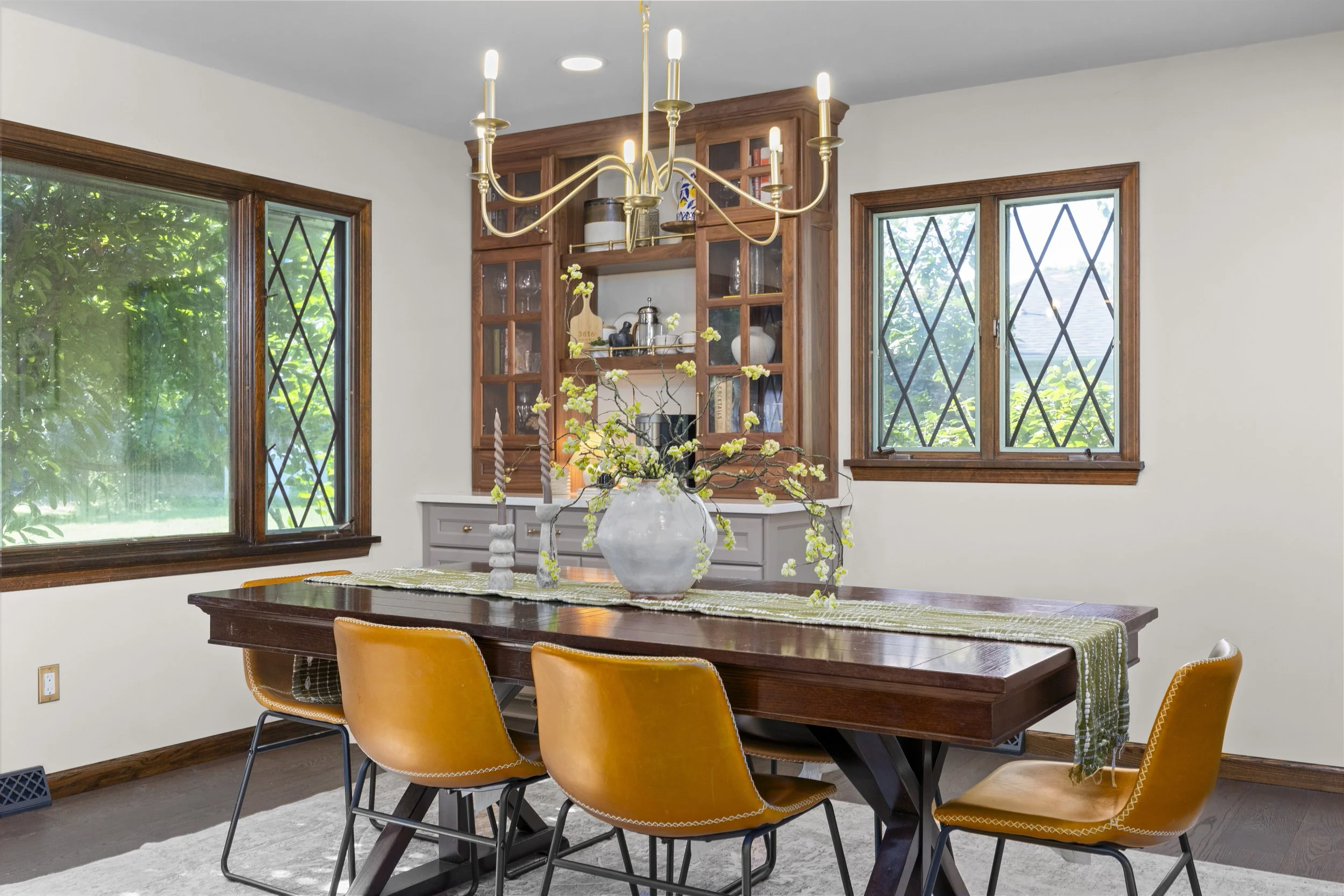 A dining room with large windows, a wooden dining table, yellow chairs, a white vase with yellow flowers, and a brass chandelier hanging from the ceiling.