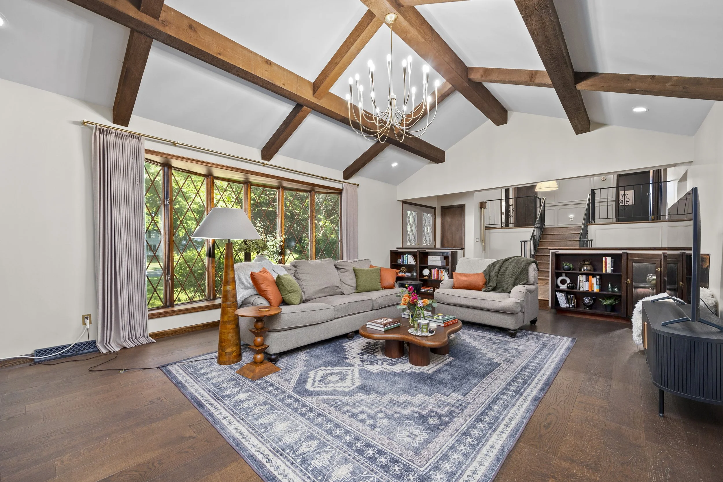 Living room with high ceiling, exposed wooden beams, a large chandelier, big windows, beige sofa with colorful cushions, a wooden coffee table with books and flowers, a lamp, and a bookshelf.