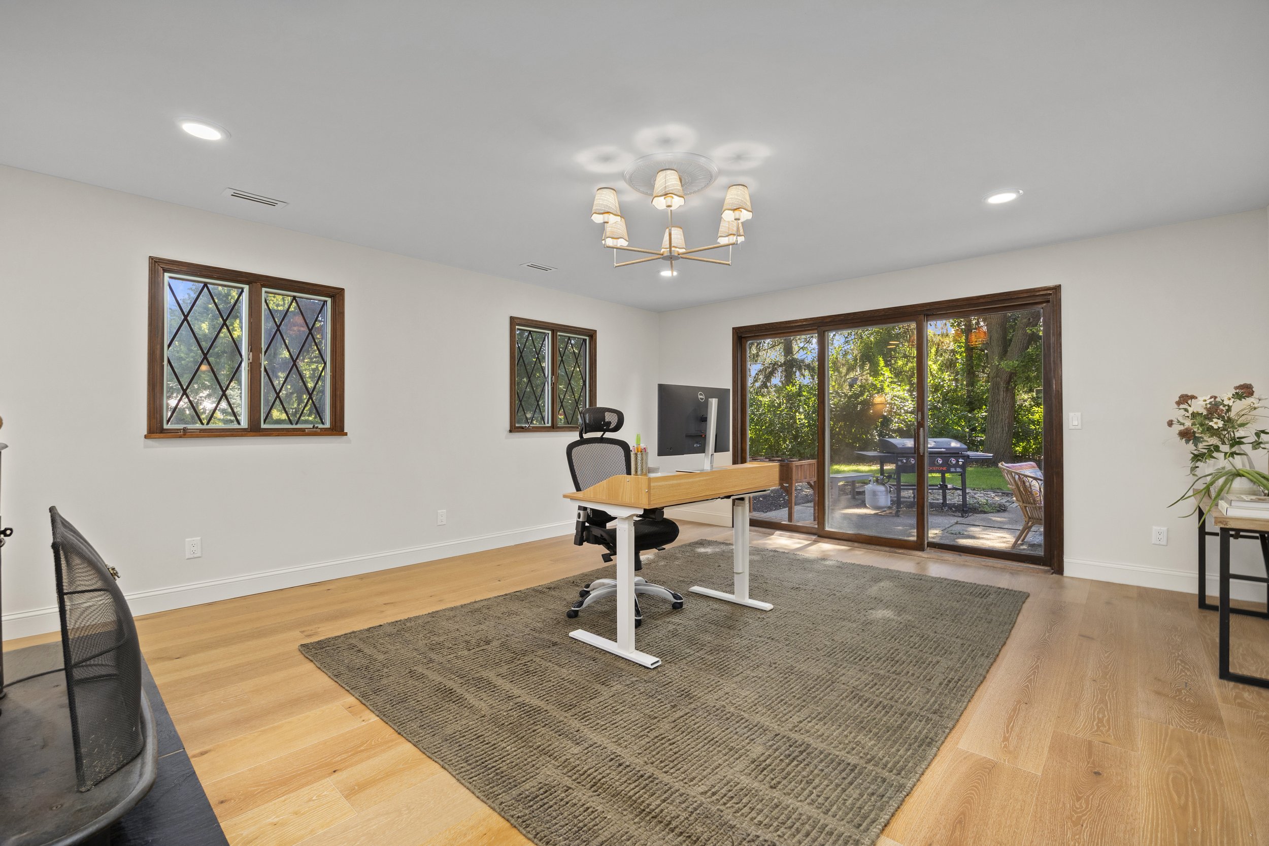 A bright home office with two windows and a sliding glass door leading outside. There is a desk with a computer, an office chair, and a beige rug on hardwood floor. Plants and outdoor furniture are visible outside.