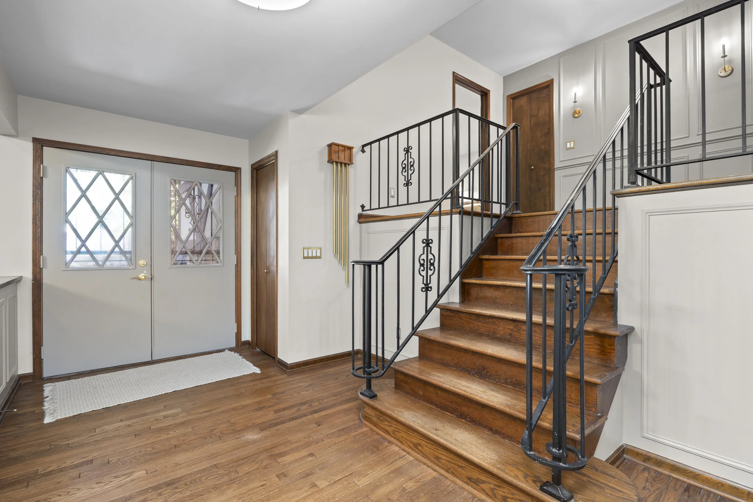 Interior of a house with wooden stairs, hardwood floor, and a white wall. There's a set of double doors with diamond-shaped windows, and a small rug in front. Wall decor includes a gold and wood coat rack or decorative piece. The stair railing is bla