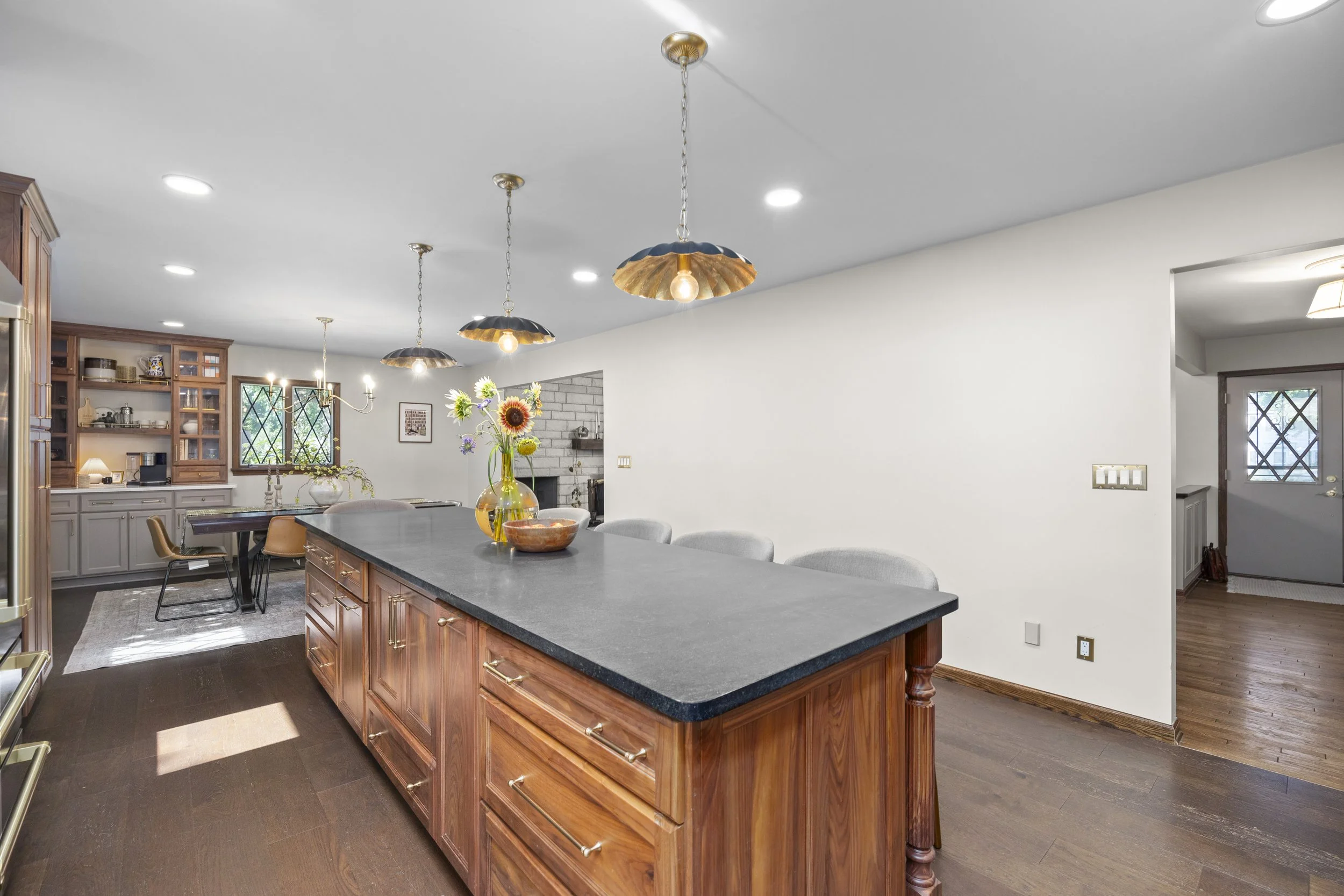 Open kitchen and dining area with a large wooden island, hanging pendant lights, a bouquet of flowers on the island, and wooden cabinets in the background.