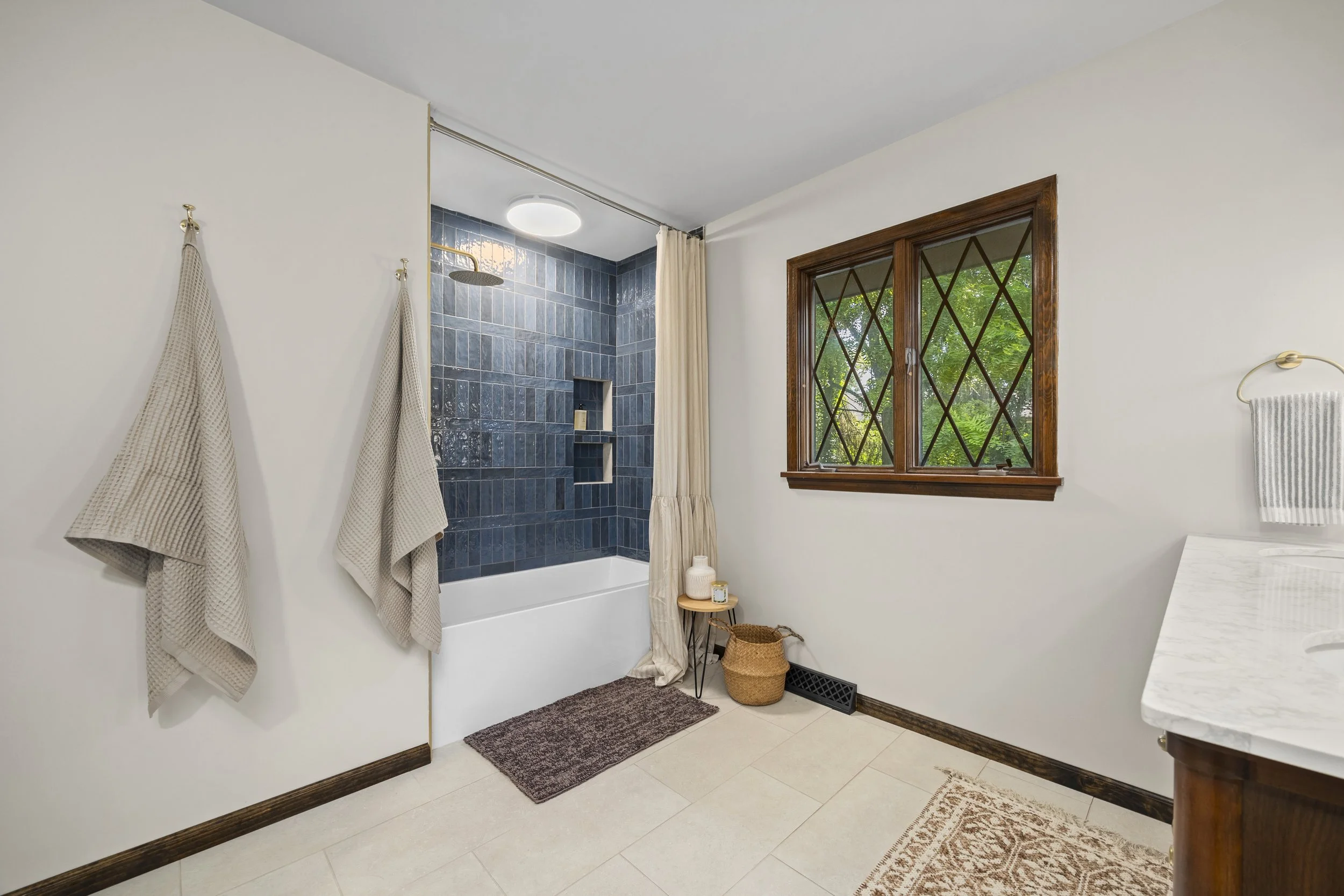 A bathroom with a blue tiled shower area, a window with wooden trim, beige towels hanging on hooks, a small basket, a brown doormat, and a white countertop with a striped towel.