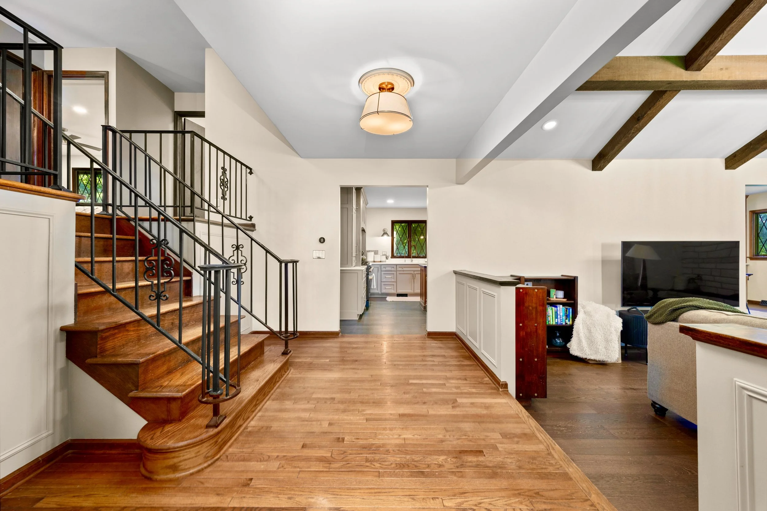 Interior view of a living room with wooden floors, a staircase with black railings, a ceiling light fixture, and a kitchen area in the background. A TV and some furniture are visible on the right.