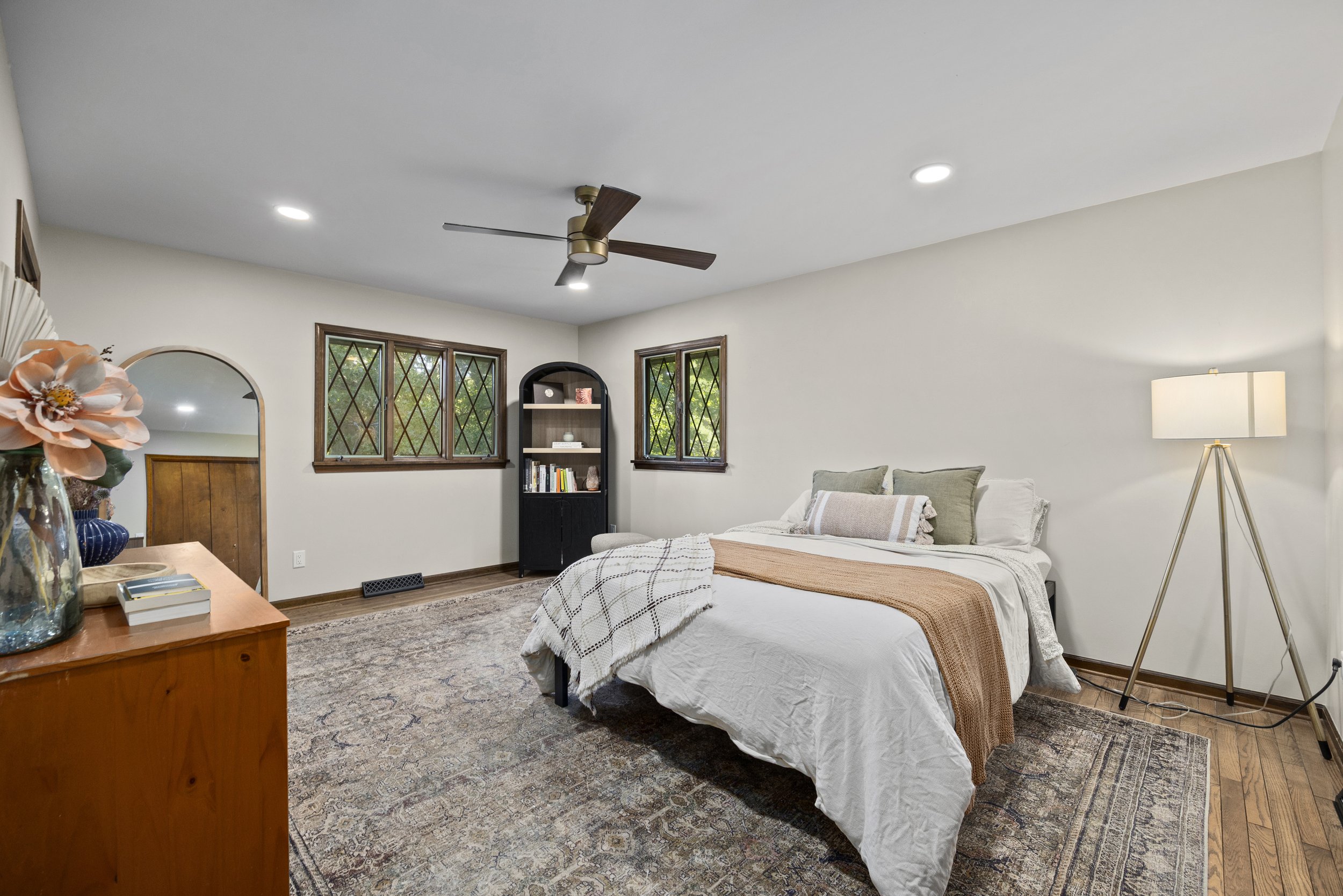 A bedroom with a white bed, pillows, and a blanket, next to a lamp, with windows, a bookshelf, and a ceiling fan.