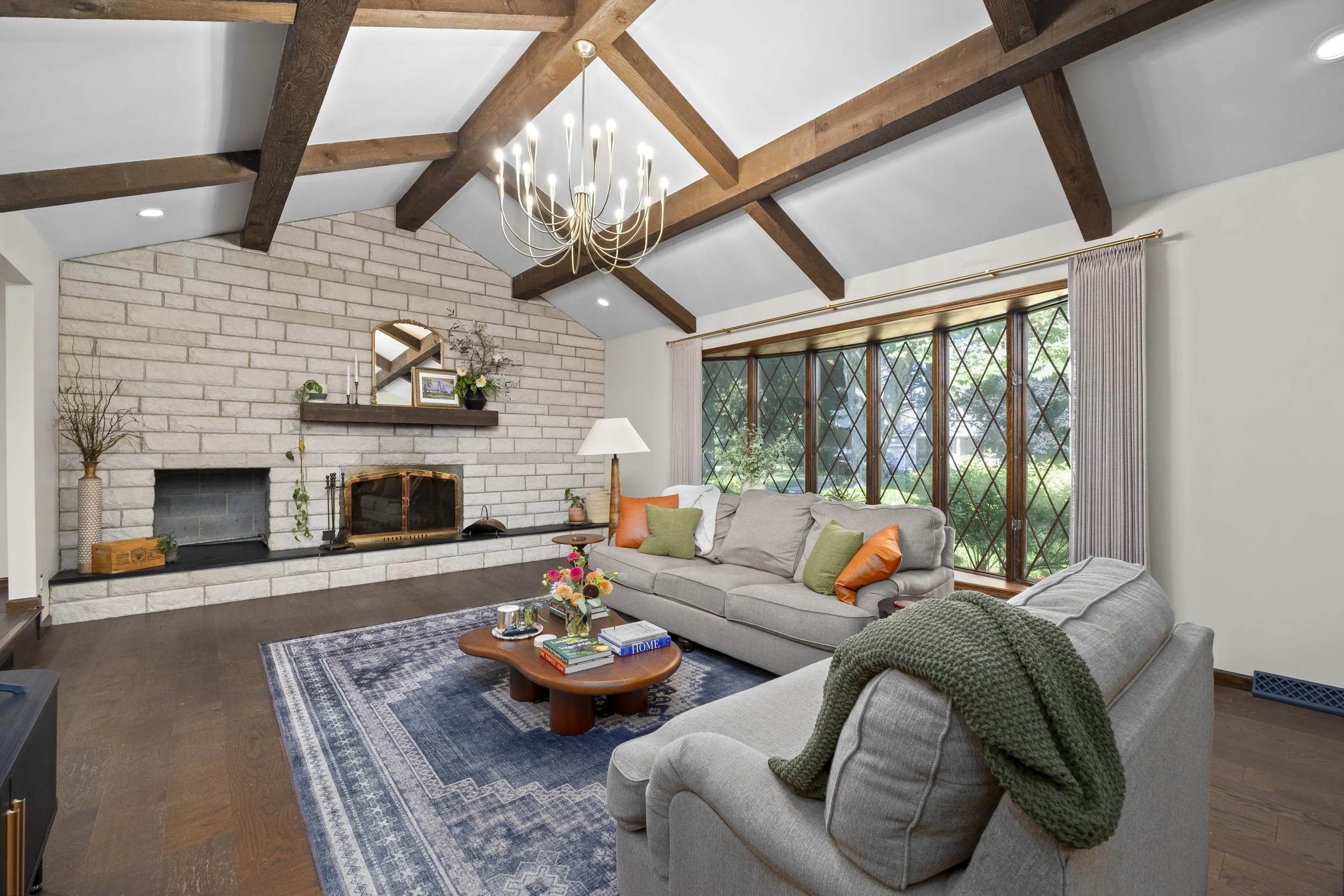 Living room with a brick fireplace, wooden beams on the ceiling, a chandelier, large windows with curtains, and a beige sofa with colorful pillows, a coffee table, and decorative plants.
