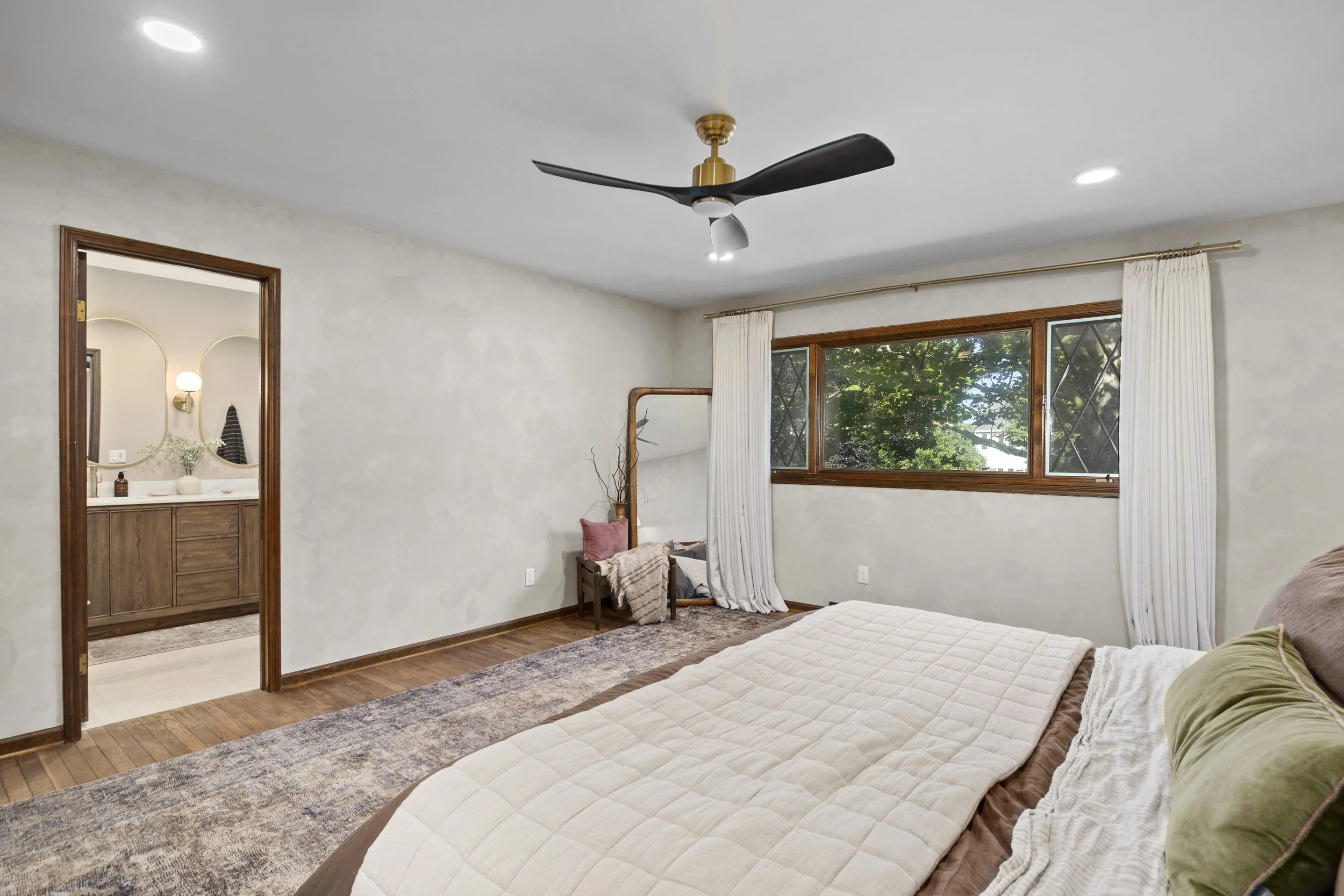 A bedroom with a large window, a bed with a quilted comforter, and a wooden chair with a pillow and blanket. The room has white walls, a ceiling fan with black blades and brass fixture, and a view of greenery outside.