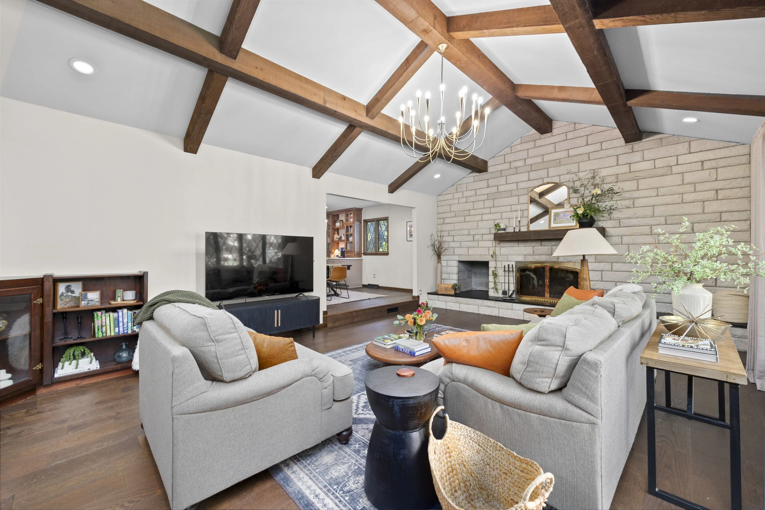Living room with vaulted ceiling, exposed wooden beams, a chandelier, a stone fireplace, and modern furniture including a sofa, armchair, and coffee table, decorated with plants and artwork.