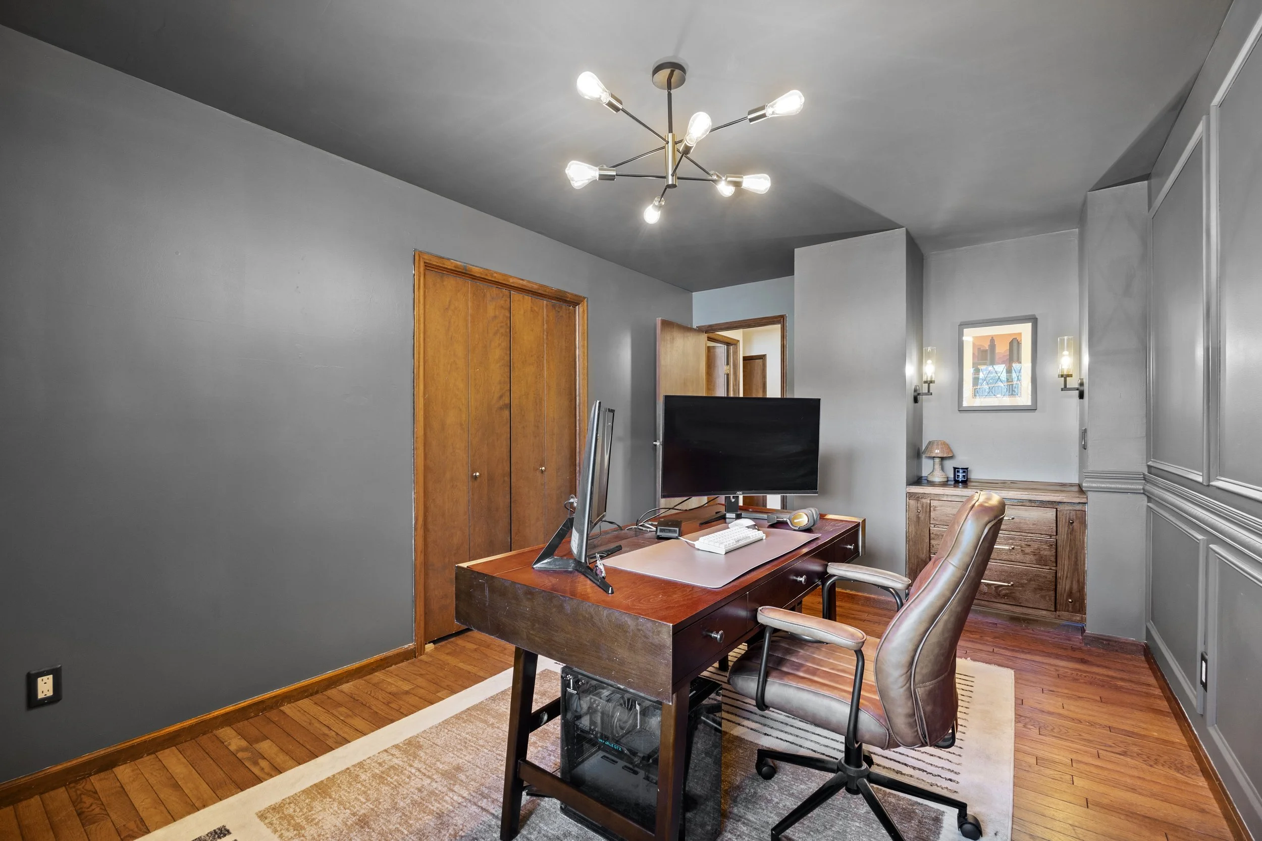 Home office with wooden desk, computer monitor, chair, and gray walls with wooden doors and a window with city skyline view.