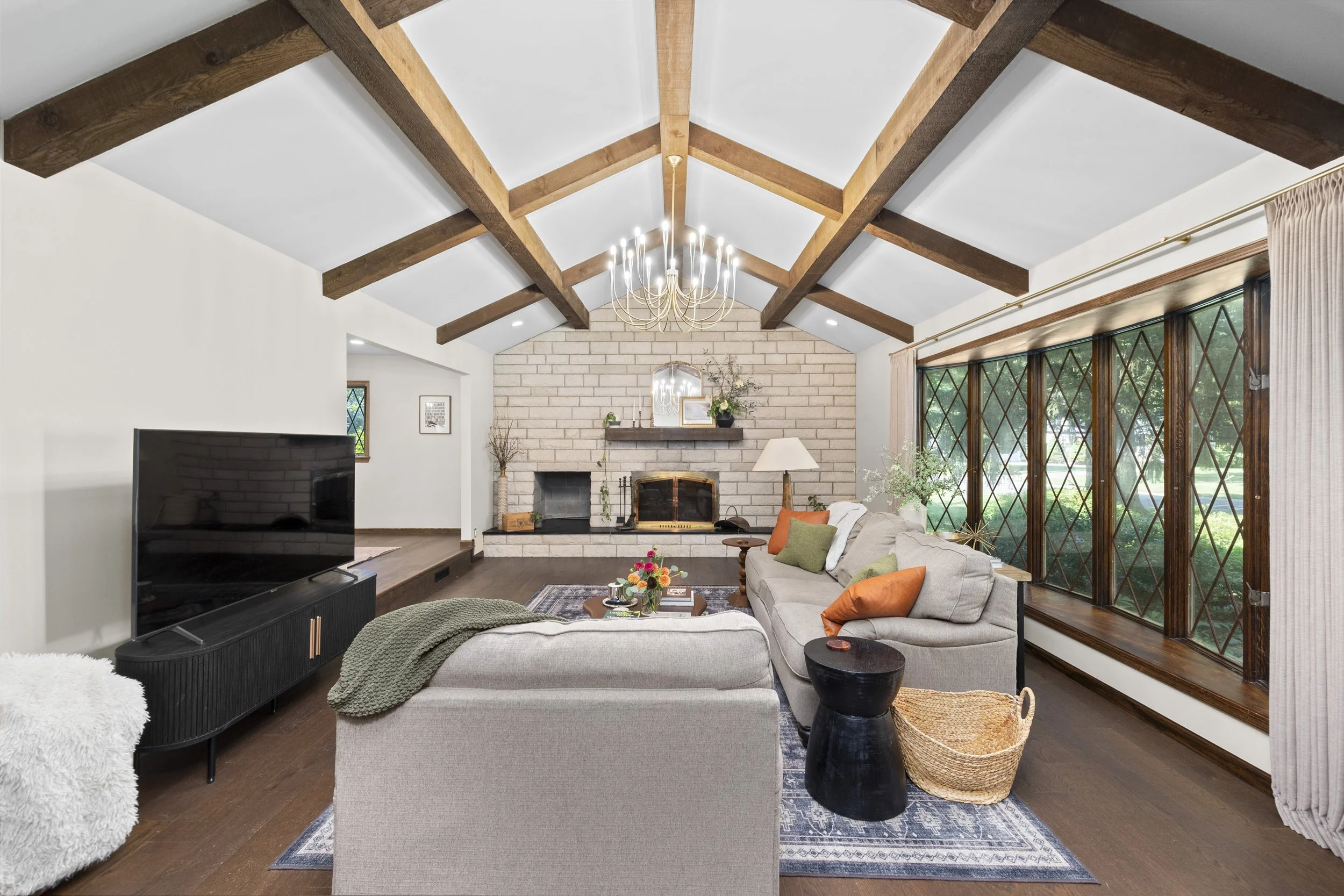 Living room with vaulted ceiling and exposed wooden beams, white brick fireplace, beige sofa with orange and green cushions, large window with diamond-pane grid, chandelier hanging from ceiling, TV on stand, and various decorative items.