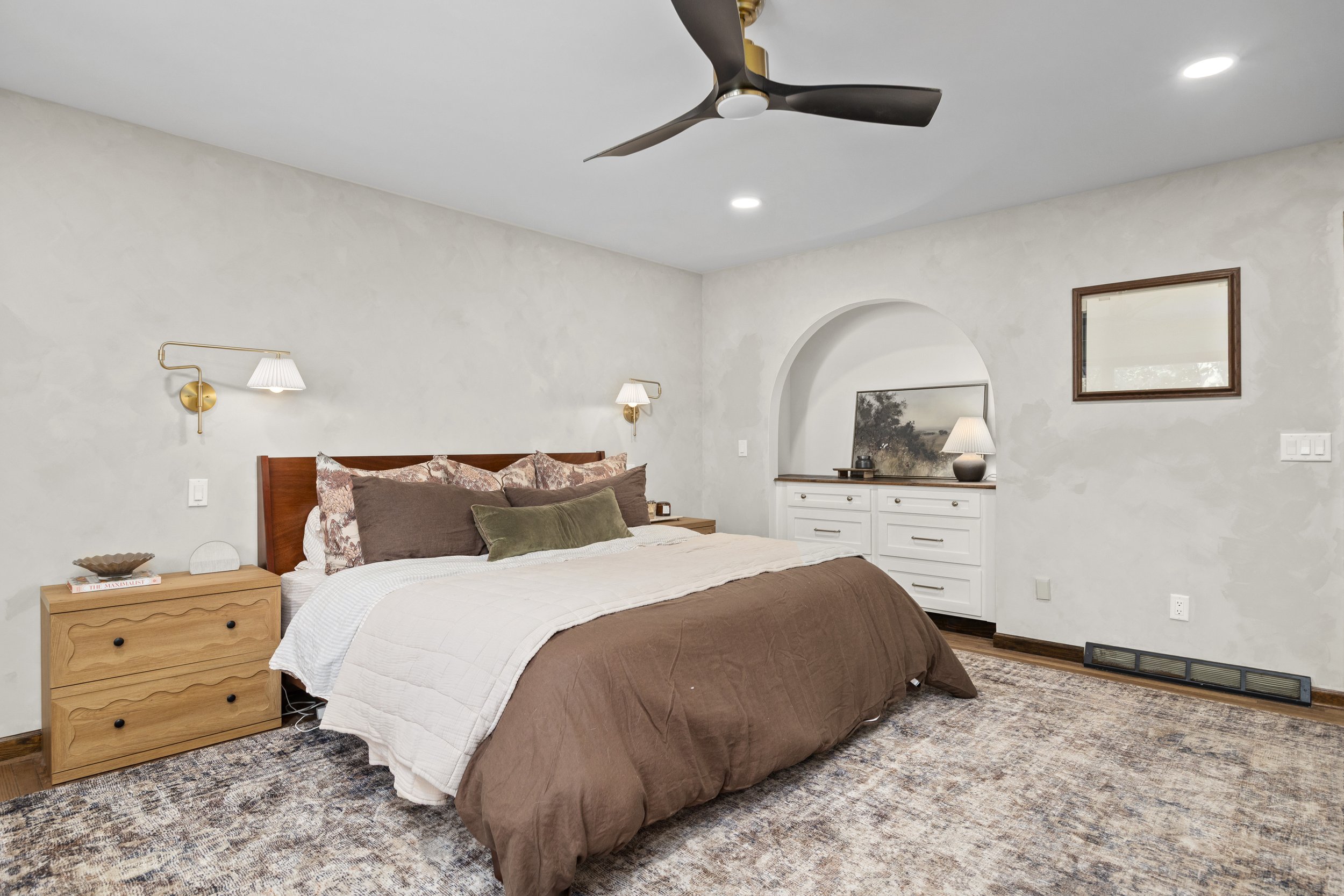 A bedroom featuring a bed with brown and white bedding, two bedside tables with lamps, a dresser with a mirror, and wall-mounted lights, with a ceiling fan and recessed lighting.