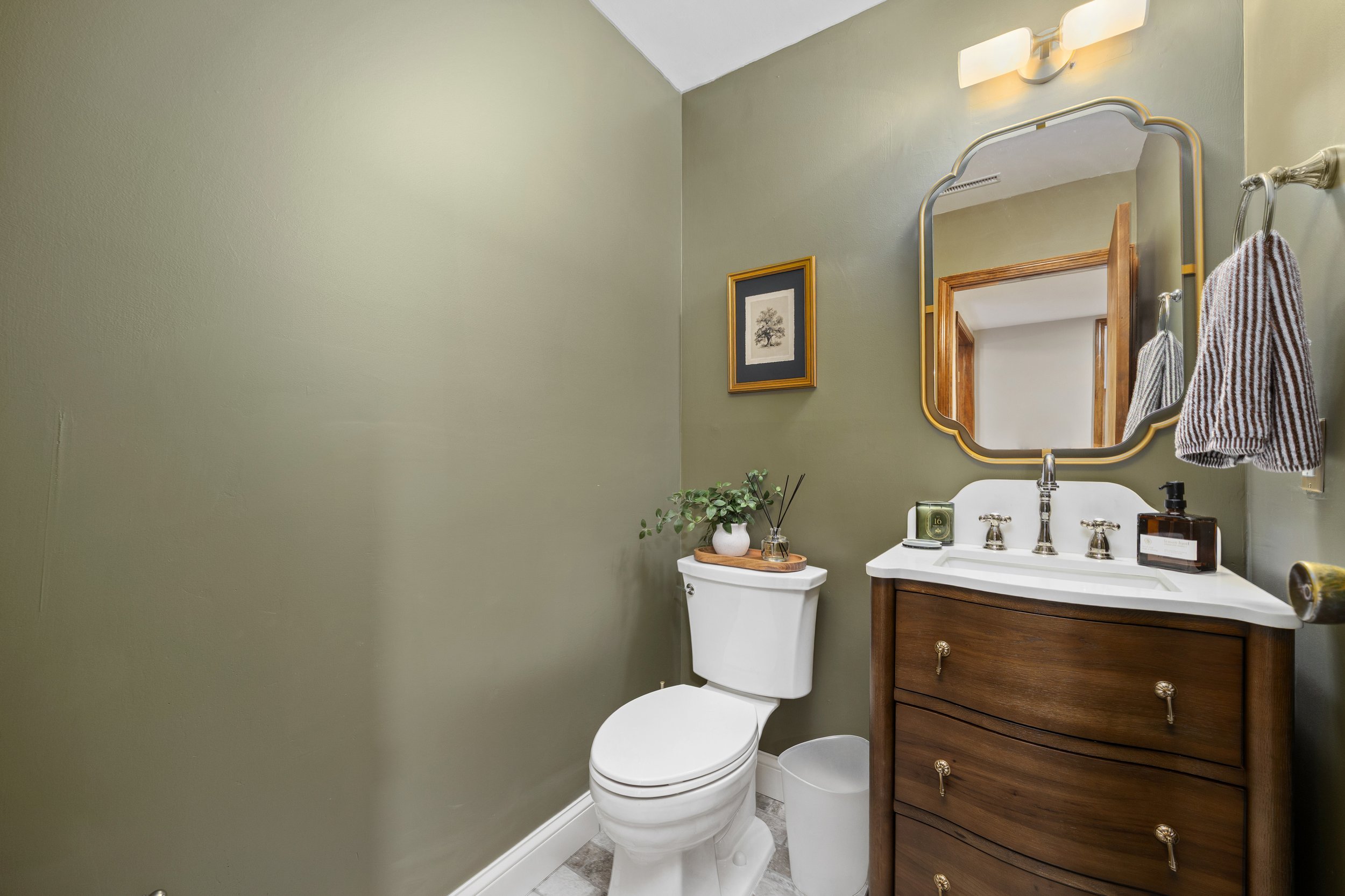 A small bathroom with a light green wall, a white toilet, a wooden vanity with a white sink, a framed black-and-white tree print on the wall, a mirror, a small potted plant, a soap dispenser, and a hand towel hanging on the wall.