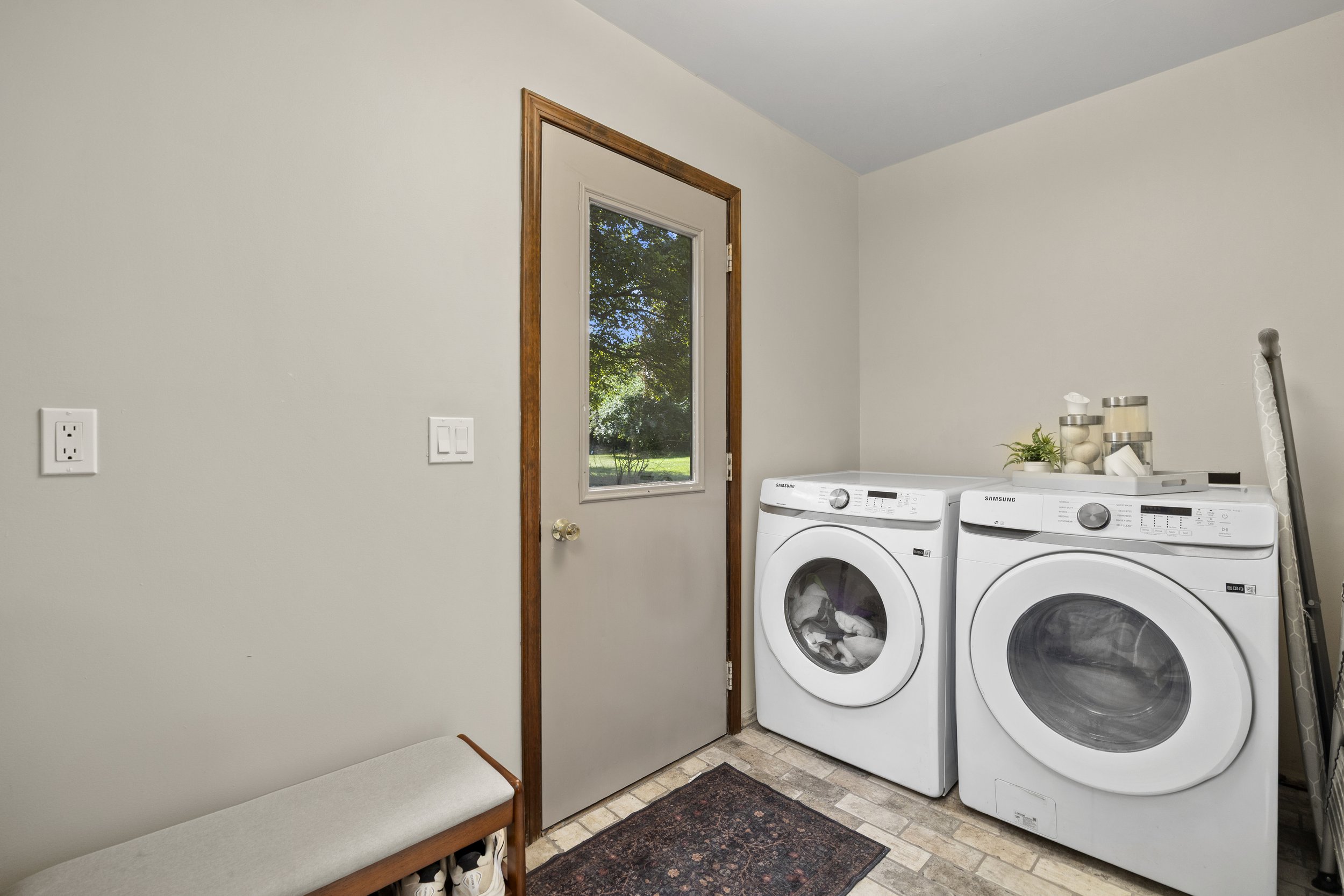 A laundry room with white walls, a window showing greenery outside, and two Samsung front-loading washing machines. There is a small shelf with decorative items, a cleaning tool leaning against the wall, and a bench with shoes underneath.