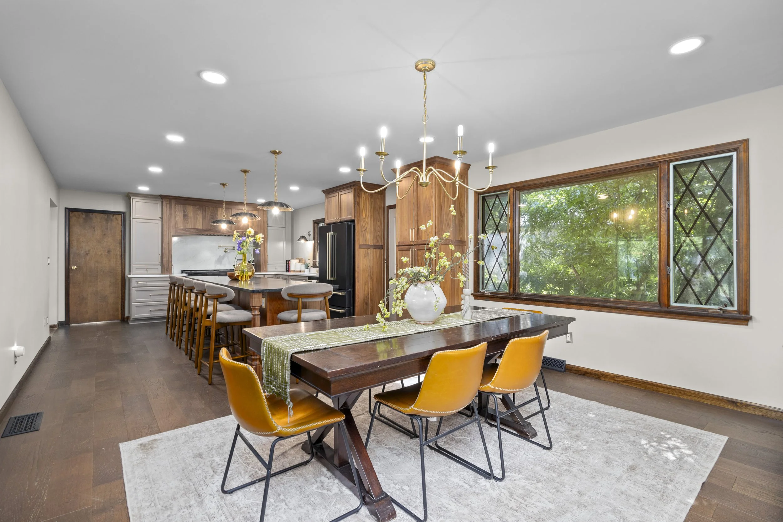 Bright kitchen and dining area with wooden cabinets, a large window, a wooden dining table with yellow chairs, a ceramic vase with flowers, and a chandelier hanging from a white ceiling.